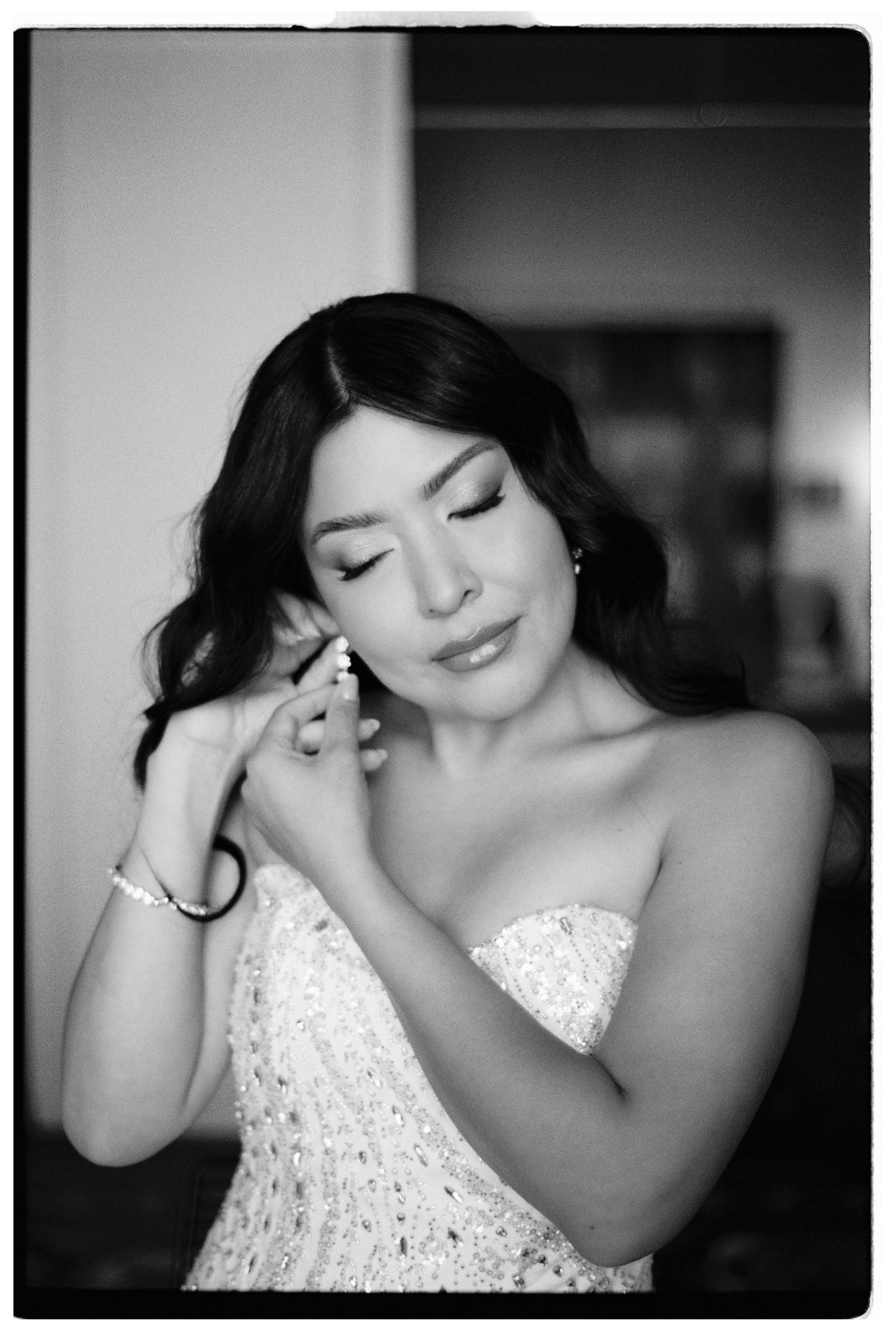 Black and white portrait of a bride putting on earrings while wearing a strapless beaded wedding dress.