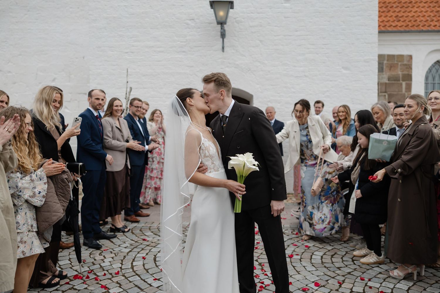 Bride and groom share a kiss outside a white church while wedding guests celebrate with confetti.