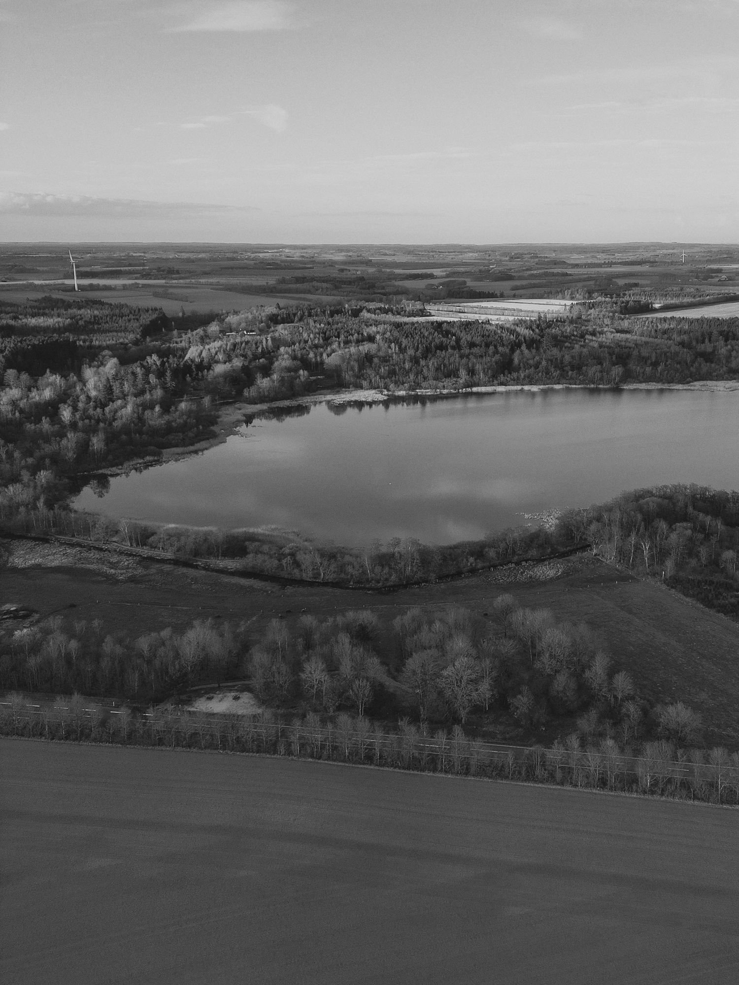 Aerial black and white photo of a serene lake surrounded by forests and farmland under a vast open sky.