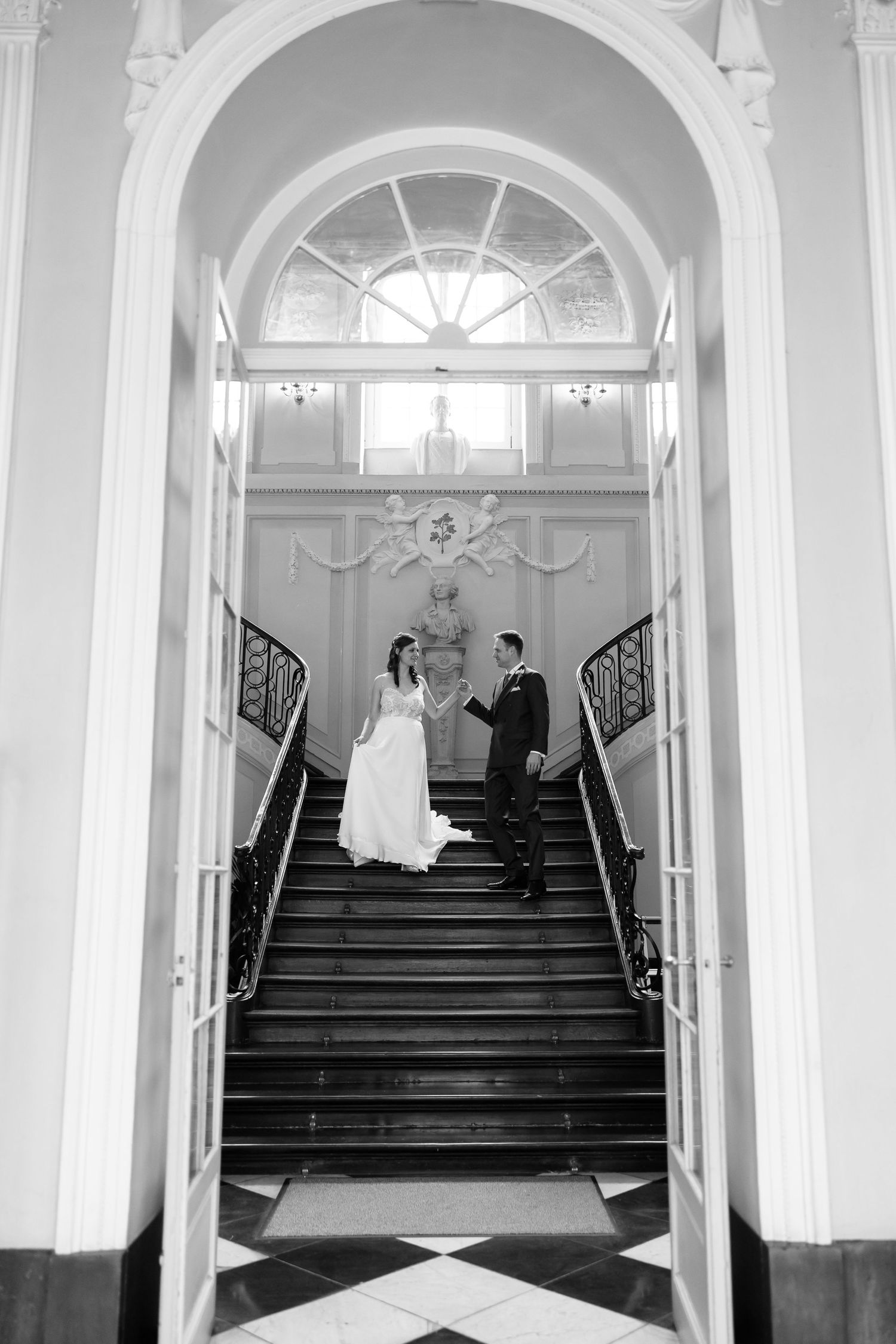 Black and white photo of bride and groom holding hands on grand staircase inside elegant wedding venue.