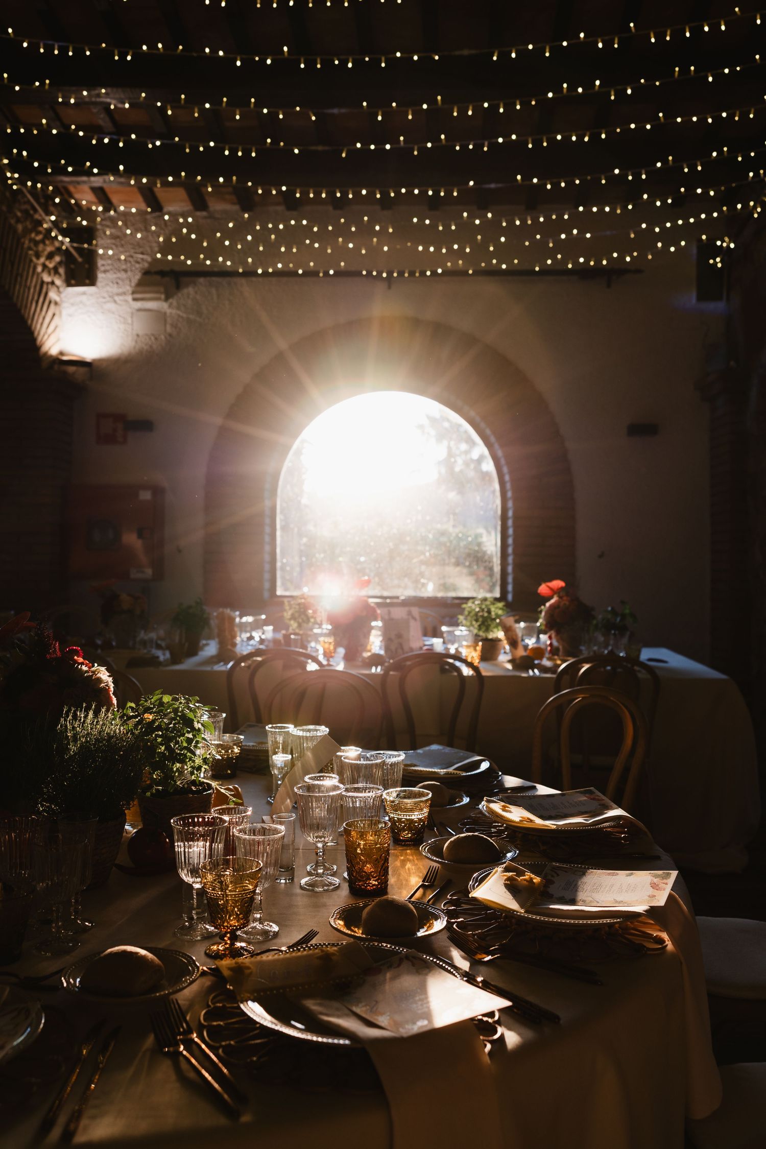 Romantic wedding reception hall with string lights, arched window, and elegantly set dining tables.