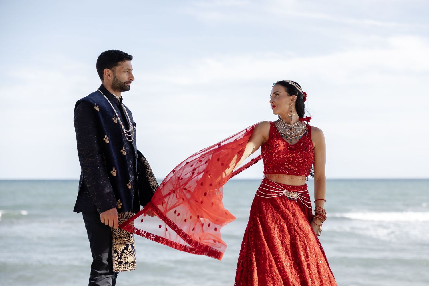 Couple in traditional Indian wedding attire posing on a beach, woman in red lehenga with flowing dupatta, man in black sherwani.
