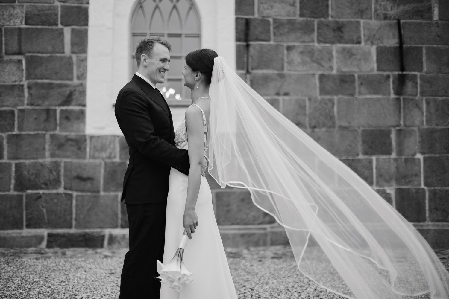 Black and white photo of a bride and groom sharing a romantic moment outside a stone church, veil flowing in the wind.