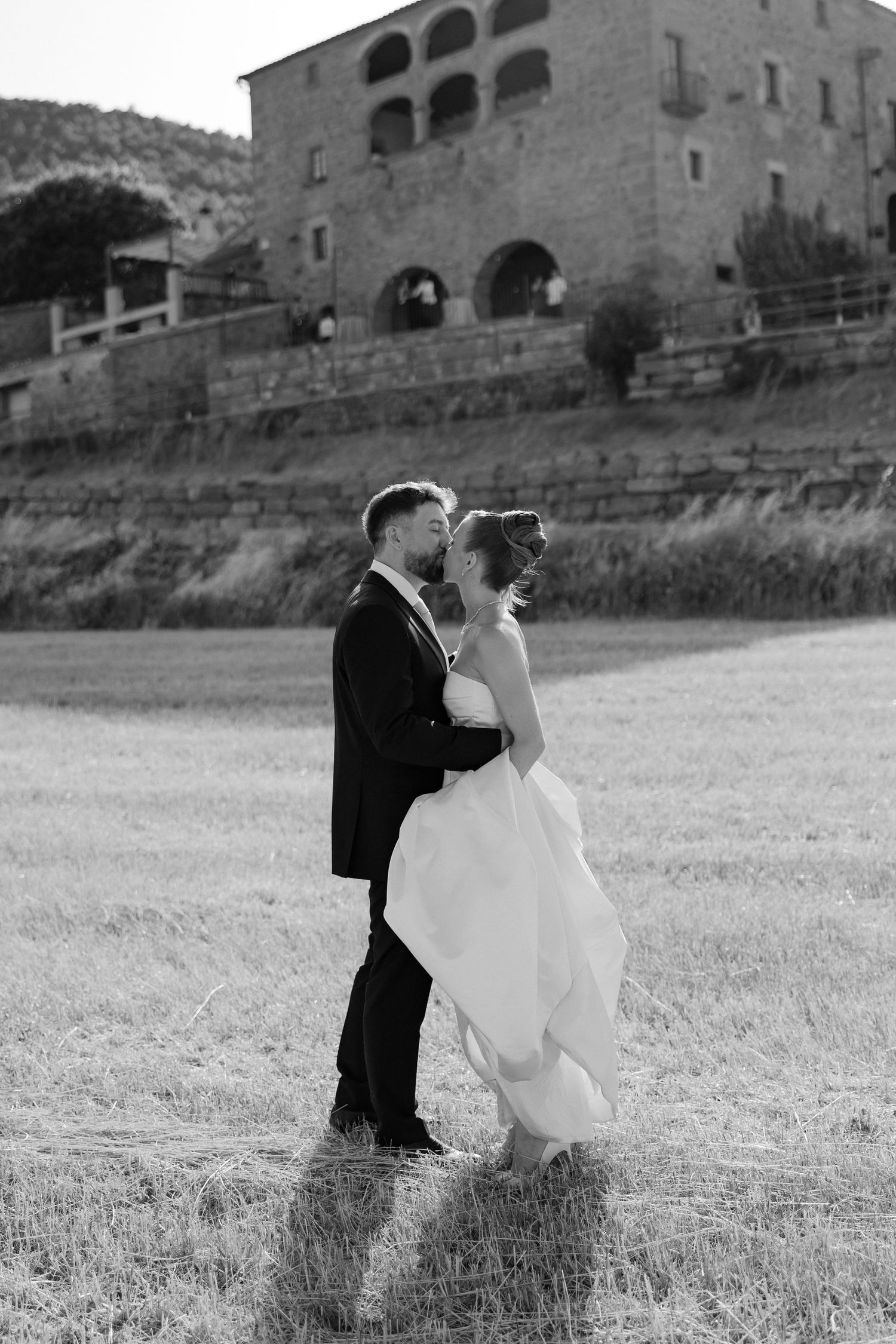 Black and white photo of a bride and groom sharing a romantic moment in a field with a stone villa in the background.