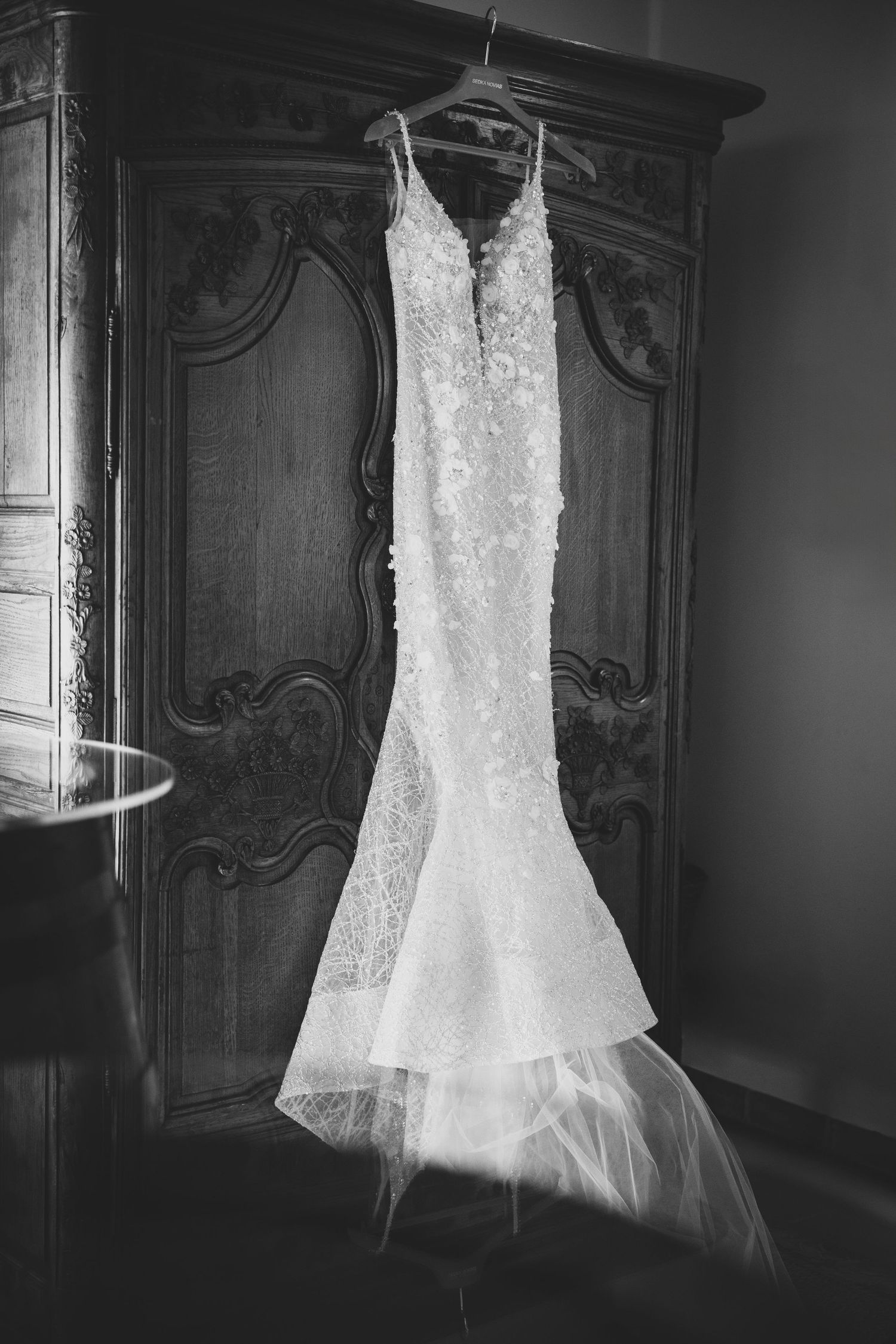 Elegant lace mermaid wedding dress hanging on an ornate wooden armoire in black and white.