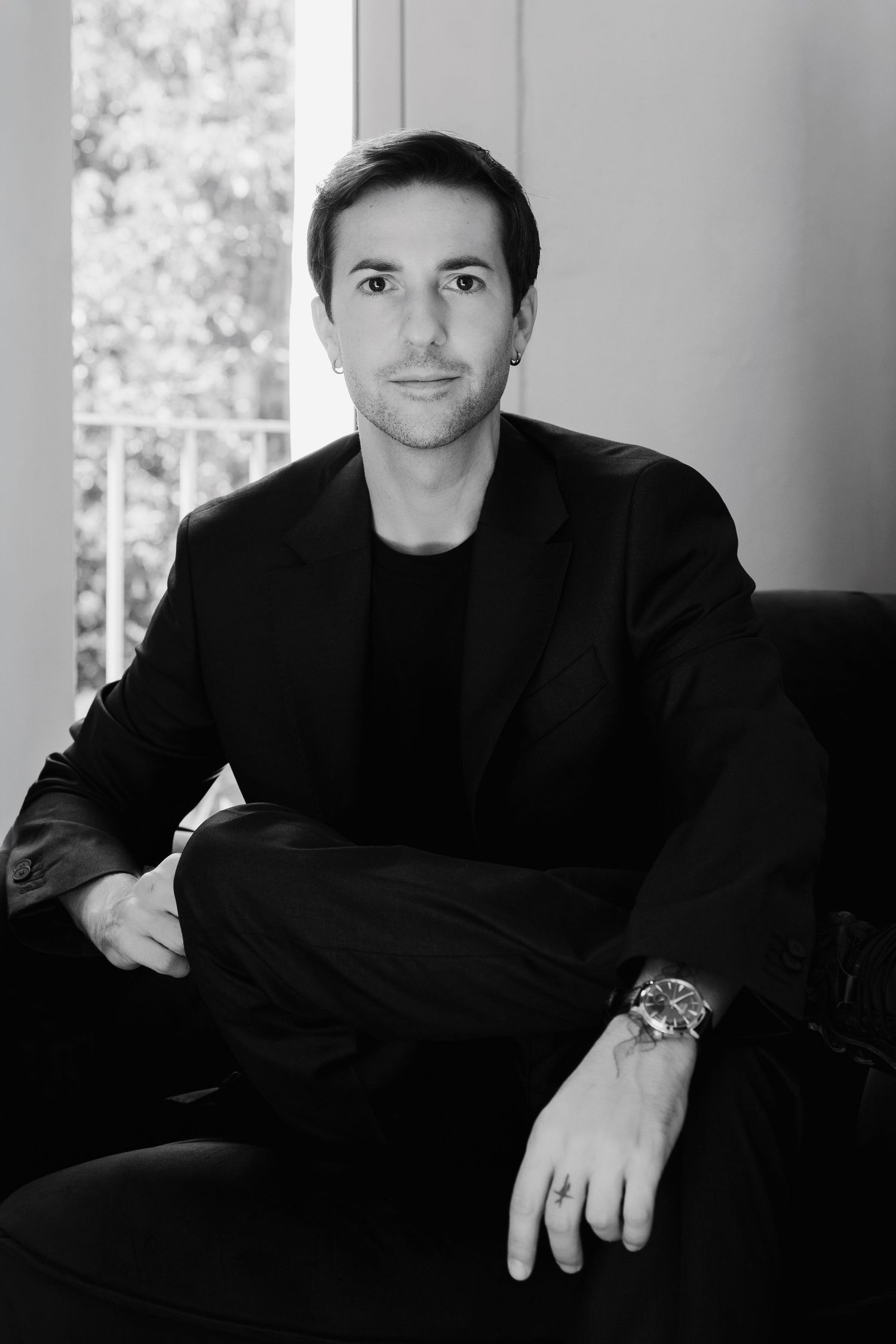 Black and white portrait of groom in black suit seated by window with watch.