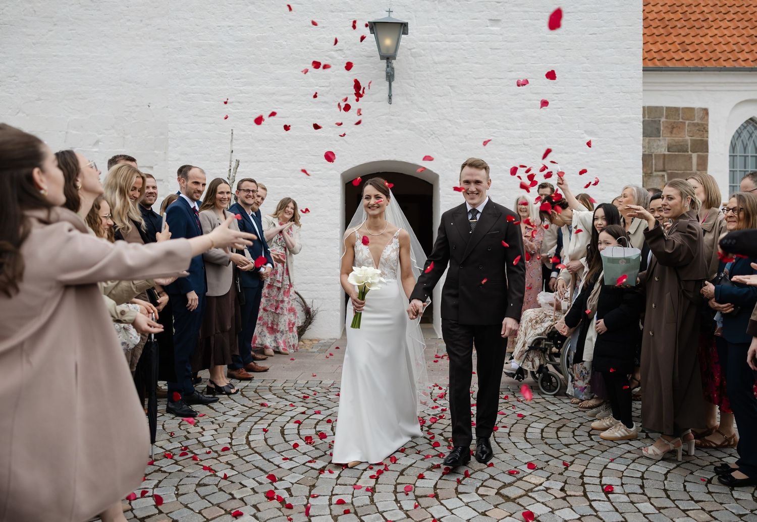 Bride and groom exit church as guests throw red rose petals in joyful outdoor wedding celebration.