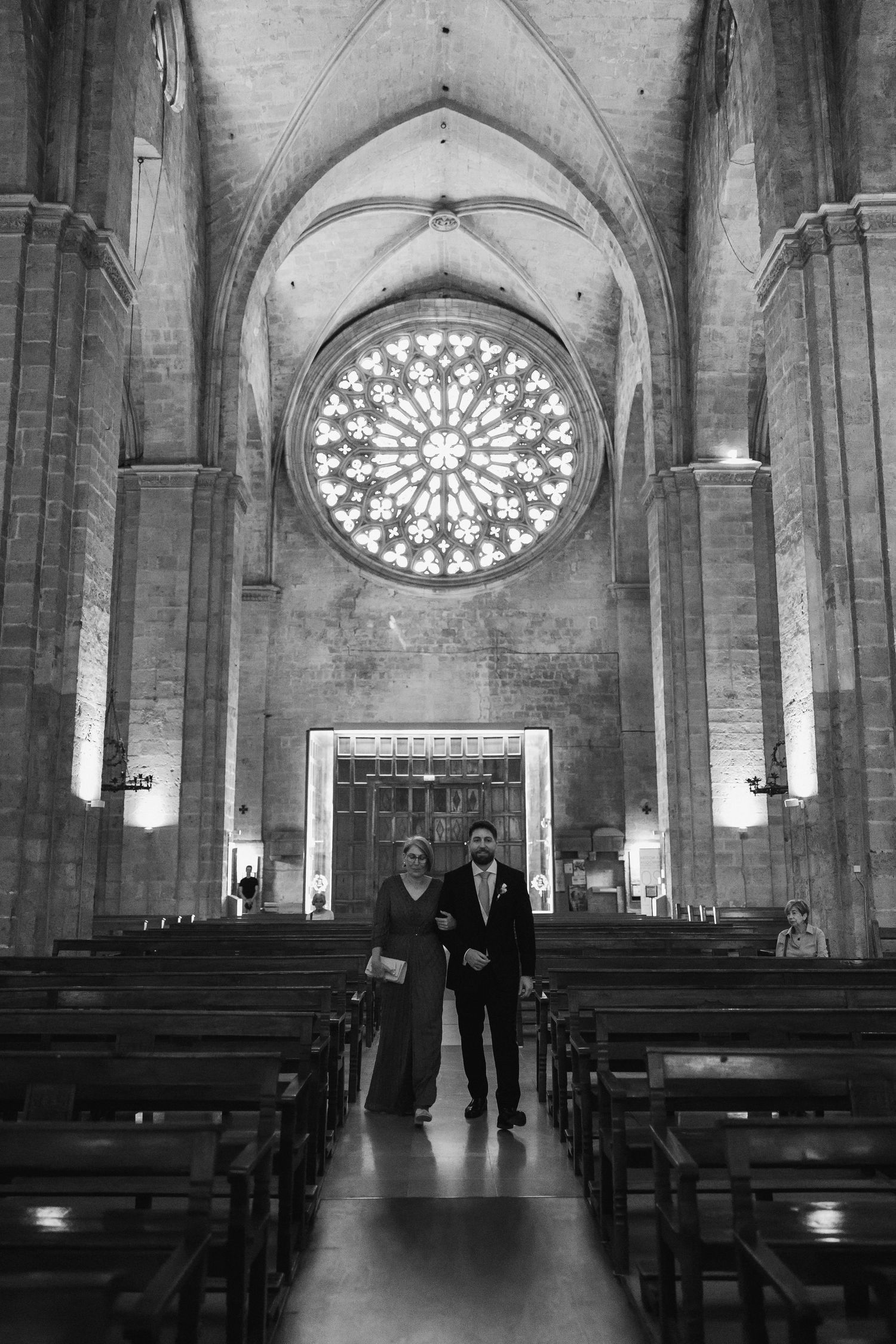 Black and white photo of a couple walking down the aisle of a grand cathedral with a stunning rose window.