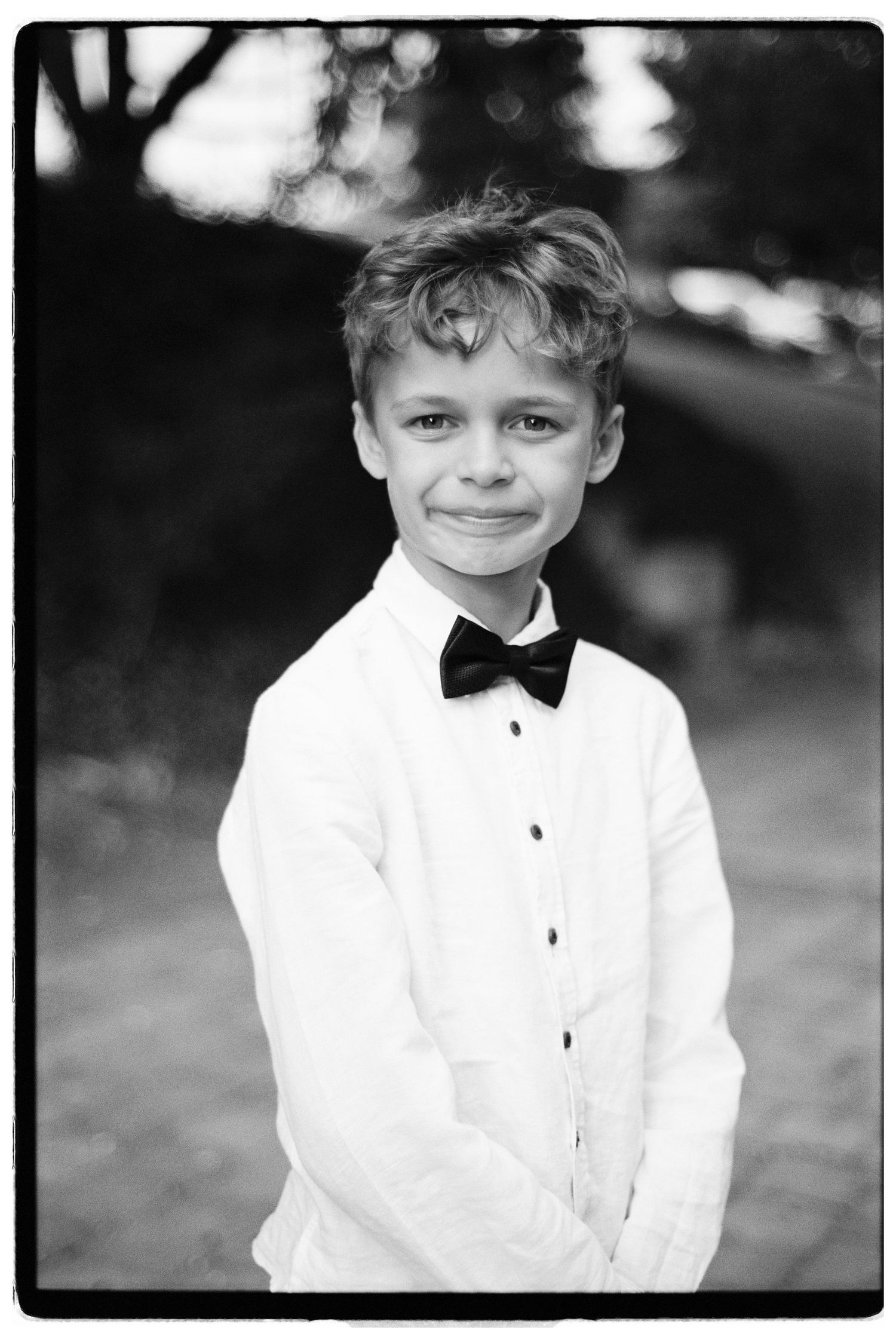 Black and white portrait of a young boy in a white dress shirt and bow tie smiling outdoors.