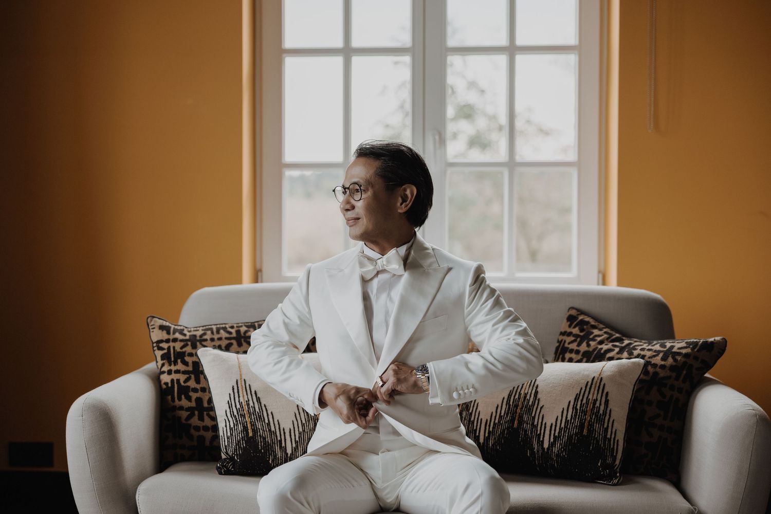 Groom in white suit with bow tie sitting on a sofa by a bright window in a golden room.