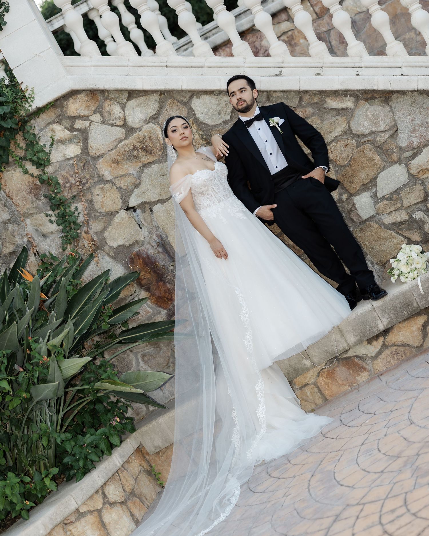 Elegant bride in off-shoulder lace wedding gown with long veil posing with groom on stone steps.