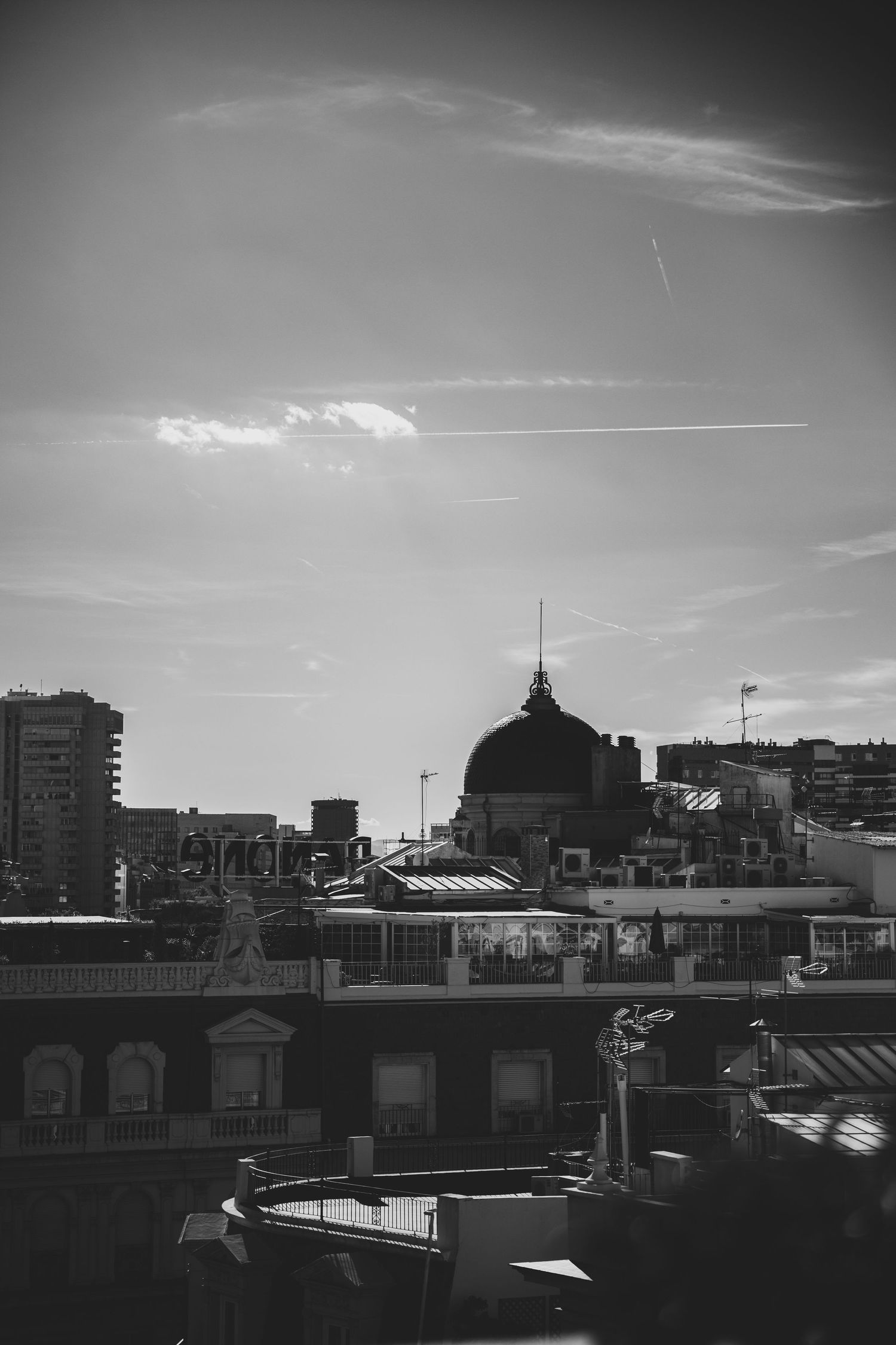 Black and white cityscape with a domed building and airplane contrail across a dramatic sky.
