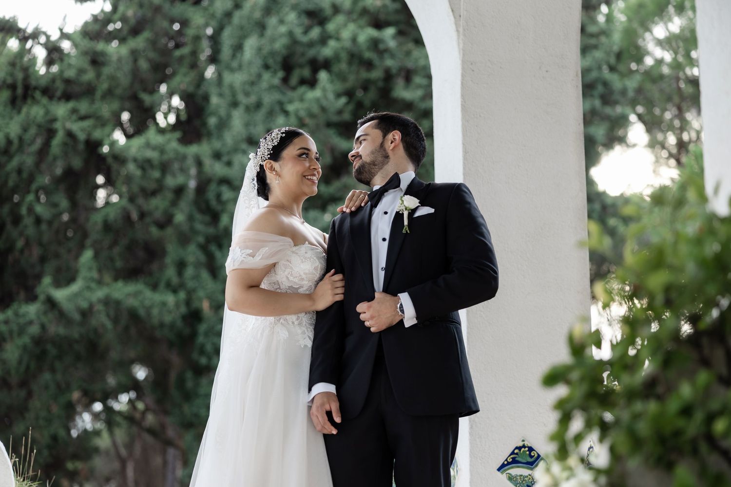 Smiling bride and groom sharing a romantic moment under white arch surrounded by greenery.