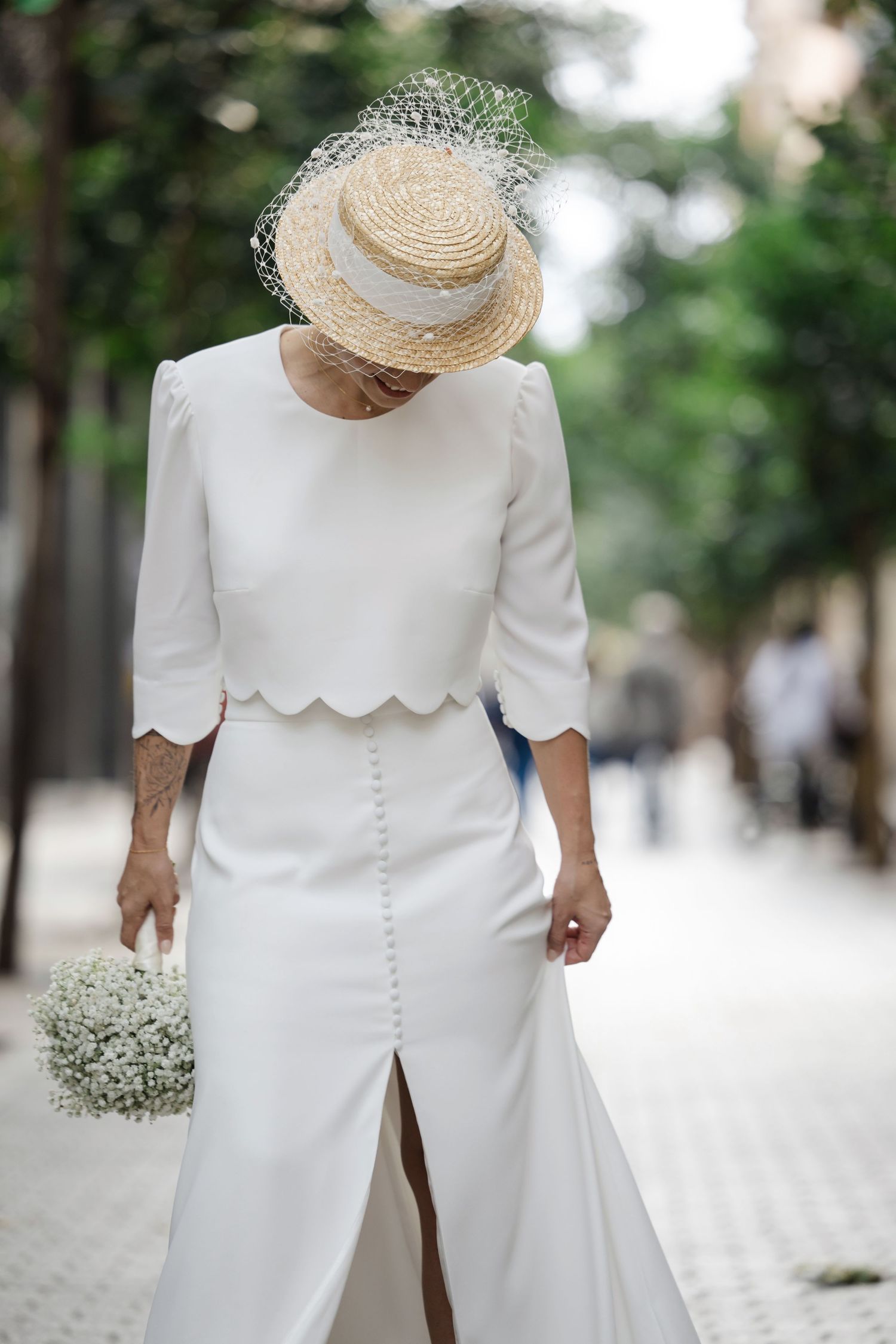 Elegant bride in white scalloped two-piece wedding dress and straw boater hat holding baby's breath bouquet.