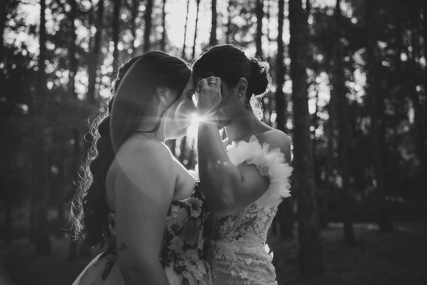 Black and white photo of two brides facing each other with sunlight flaring between them in forest.
