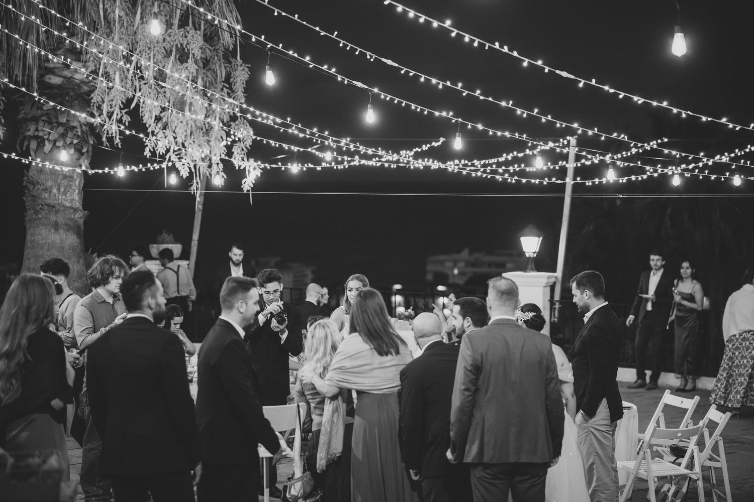 Black and white photo of wedding guests mingling outdoors at night under festive string lights in trees.