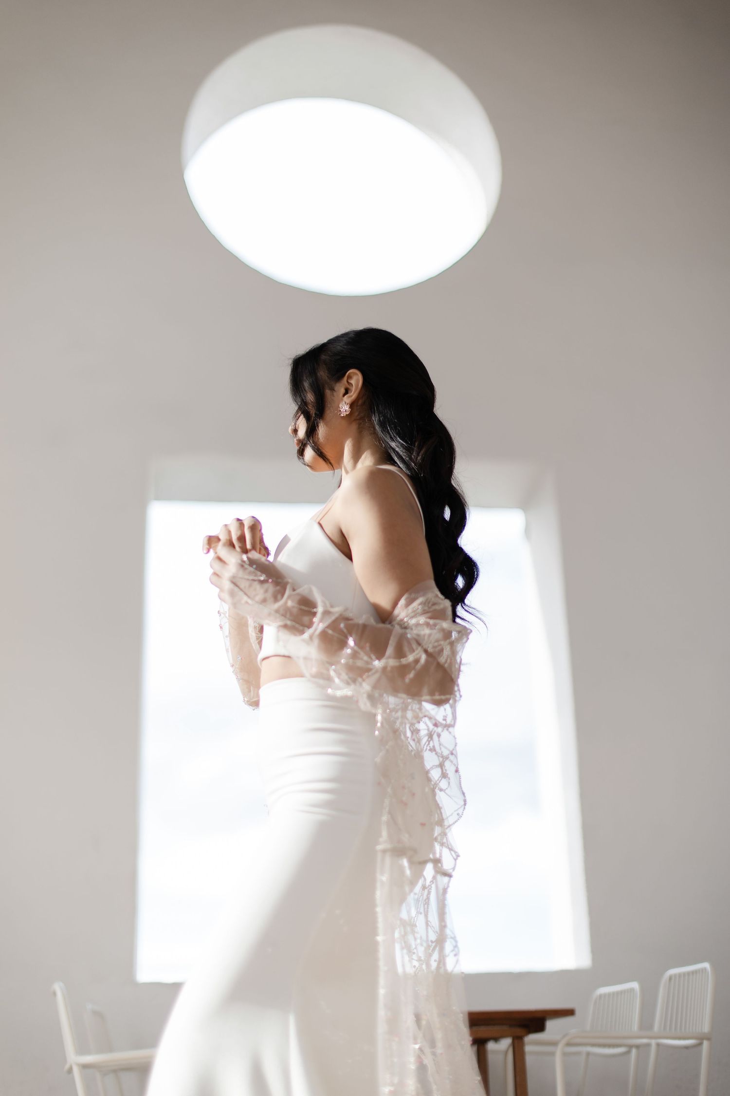 Bride with dark wavy hair wearing white skirt and lace wrap posing by bright window.