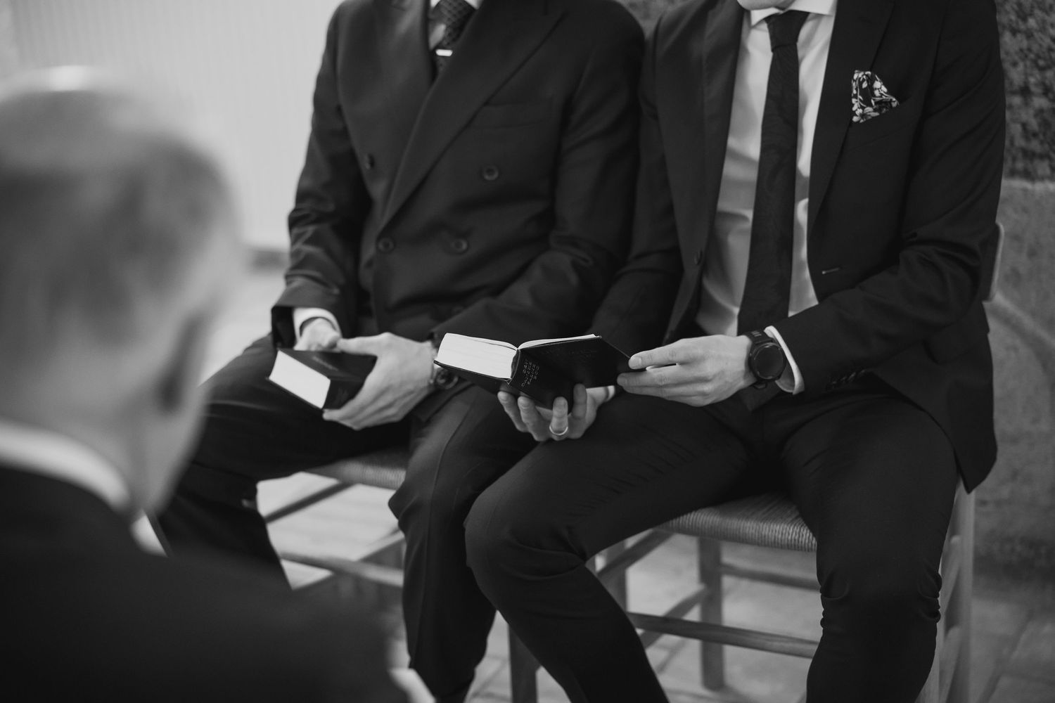Three men in dark suits holding books seated together at a formal wedding ceremony in black and white.