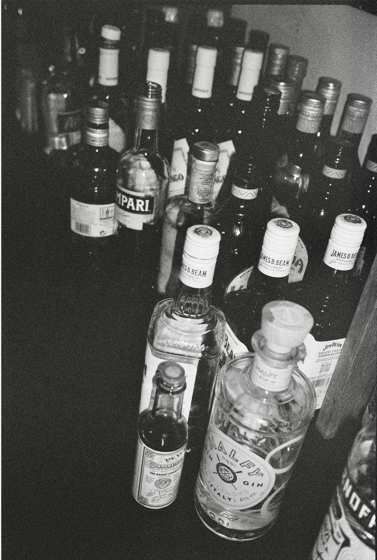 Black and white photo of assorted liquor bottles including Campari, James Beam, and gin on a bar.