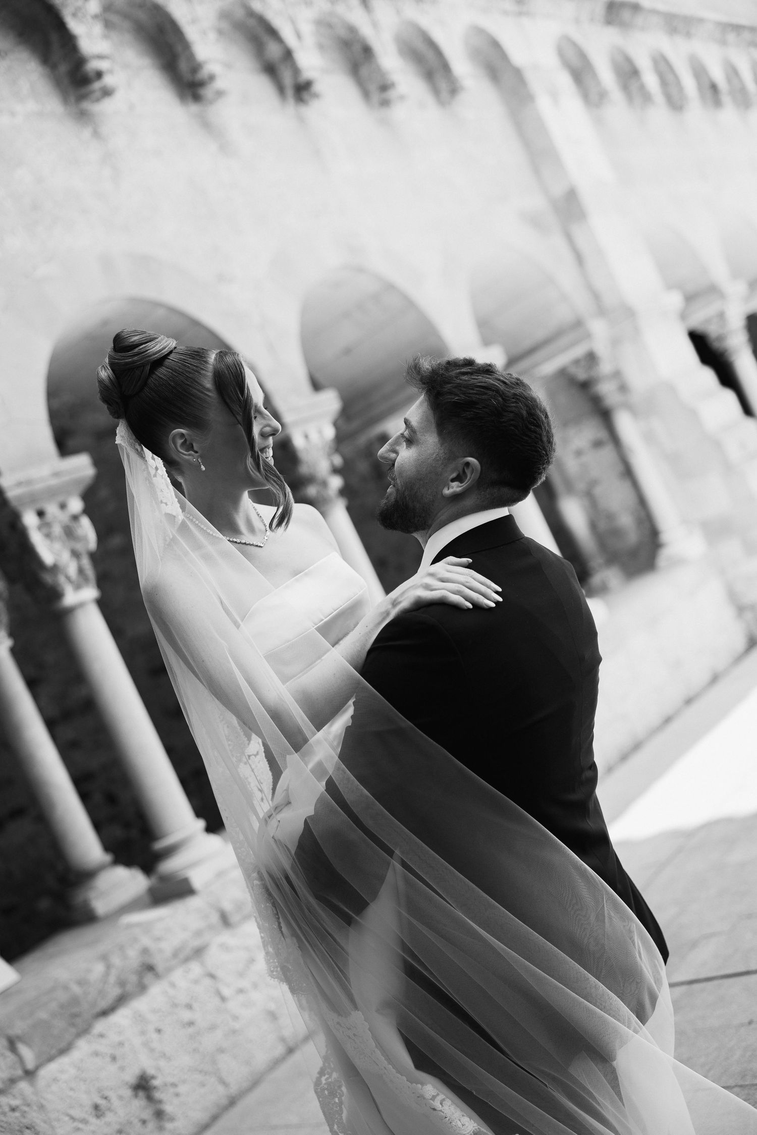 Black and white photo of bride and groom embracing, veil flowing around them in historic stone courtyard.