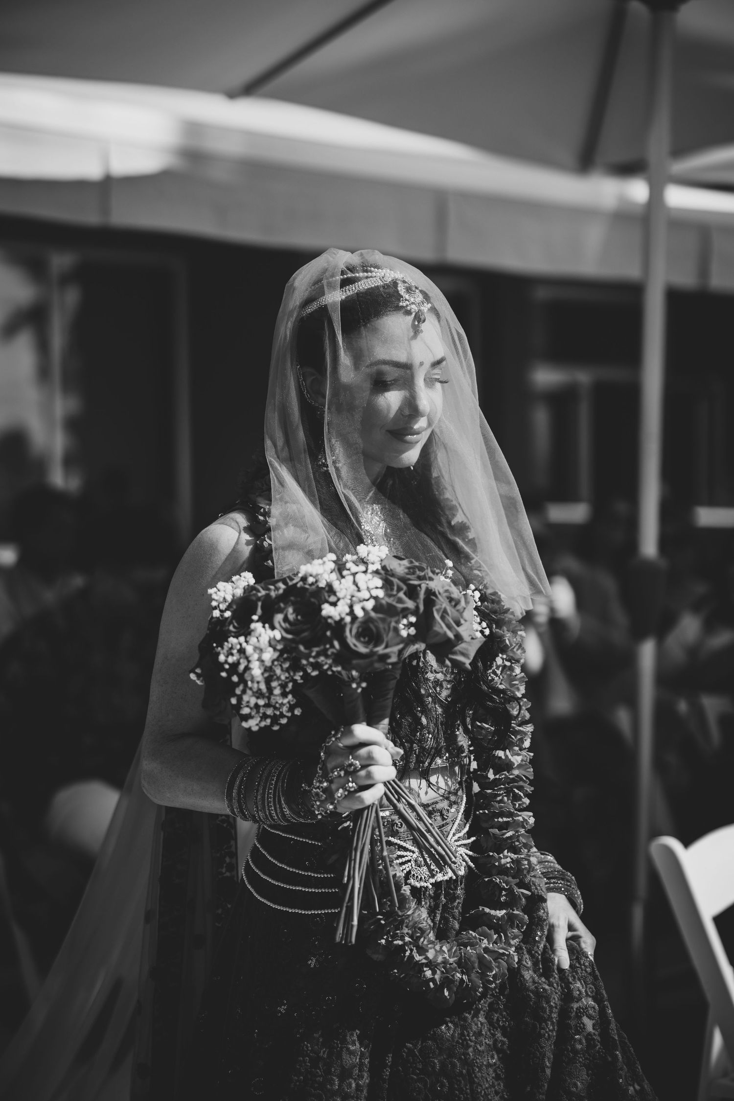 Black and white photo of a bride in a dark floral gown and veil holding a bouquet at an outdoor wedding.