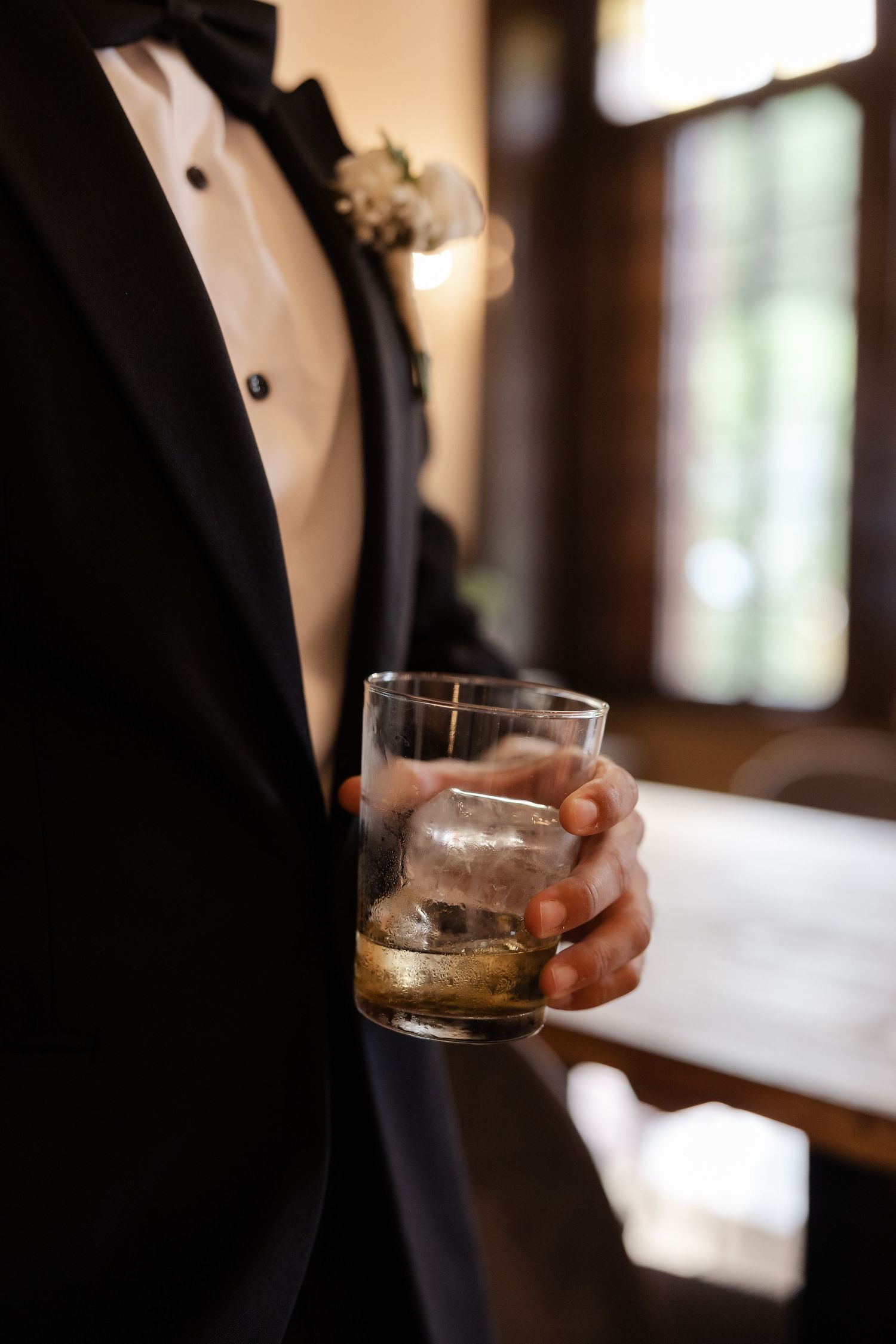 Groom in black tuxedo holding glass of whiskey with floral boutonniere at wedding reception.