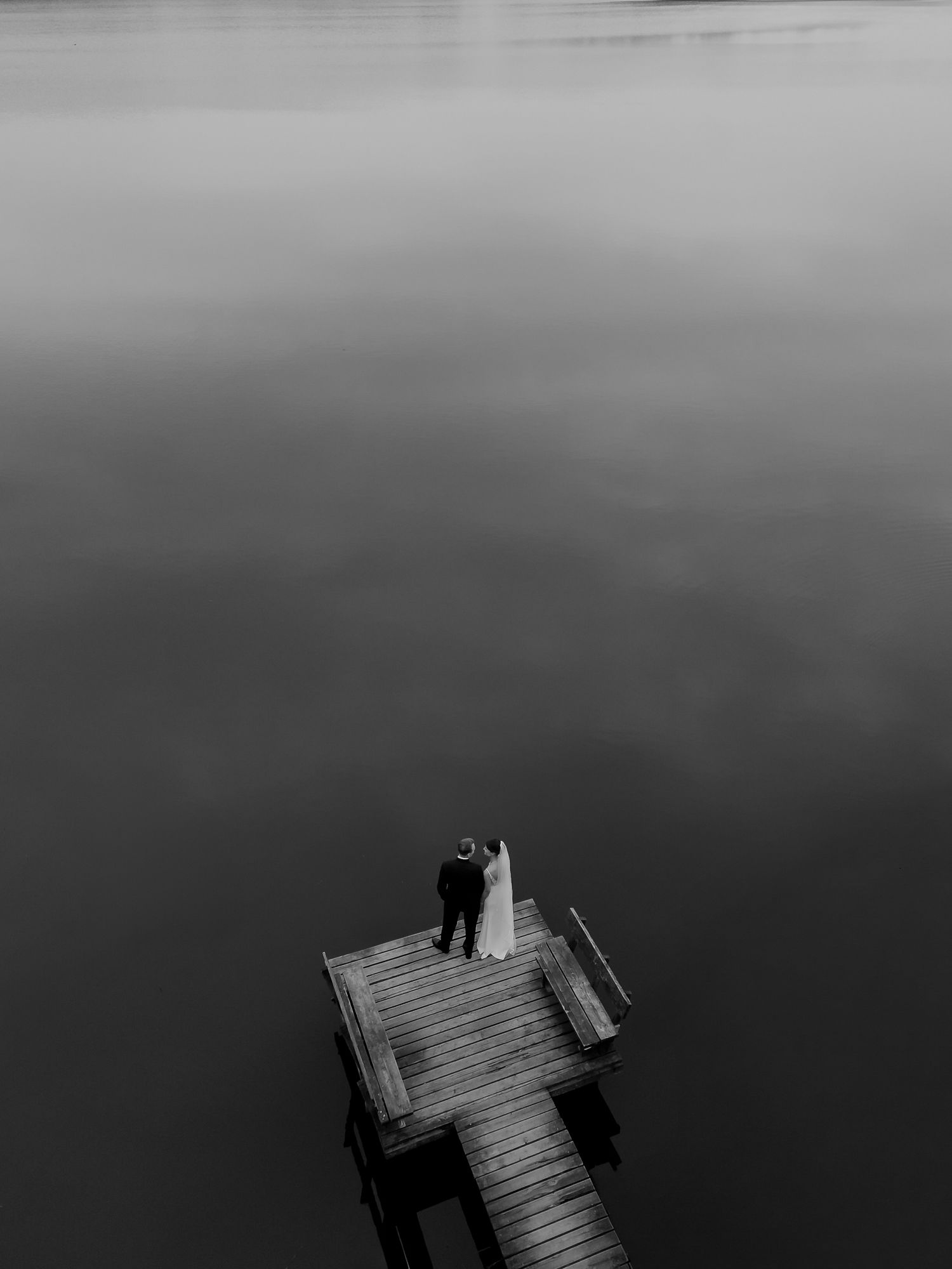Black and white aerial photo of newlyweds standing on wooden pier over tranquil lake.