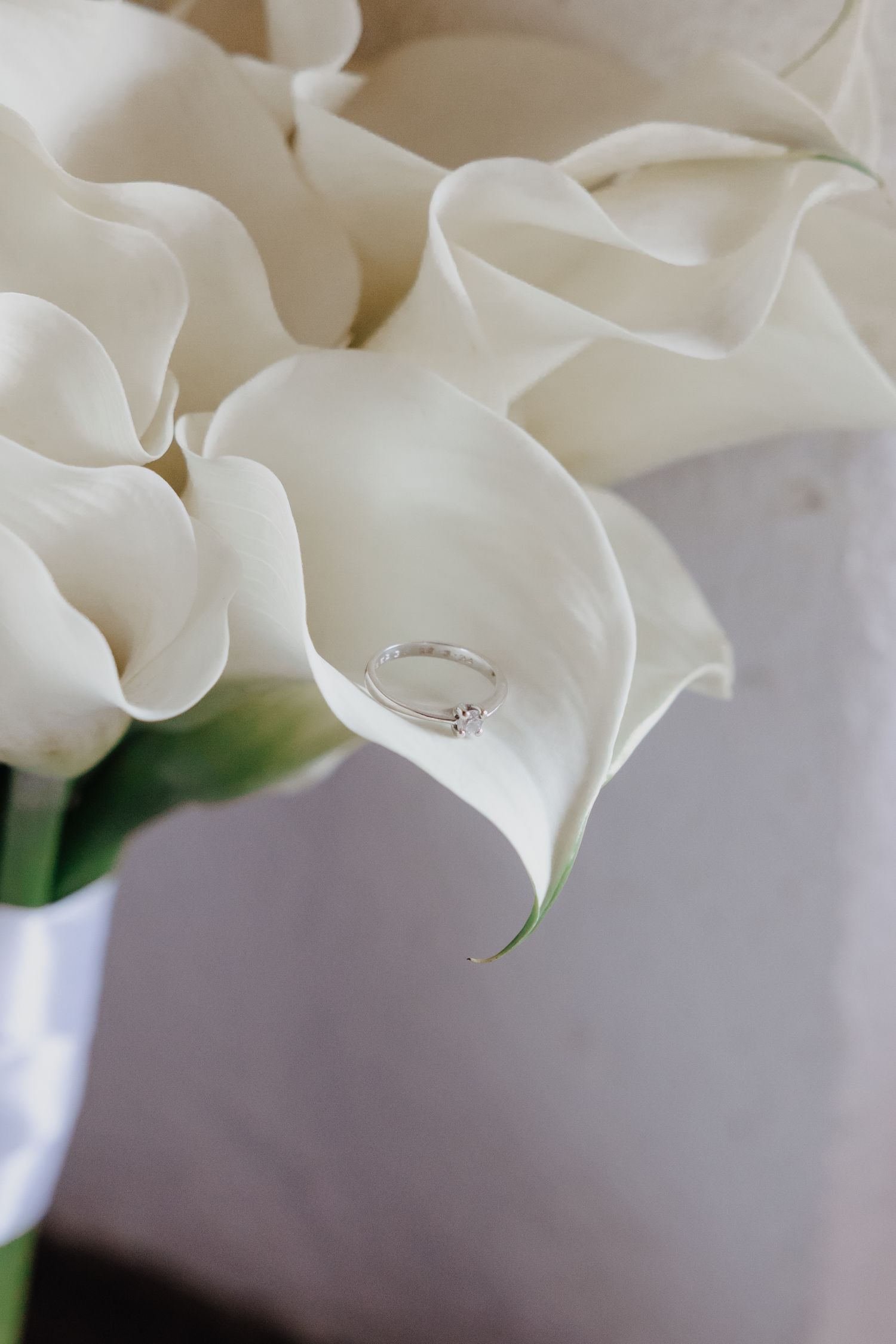 Diamond solitaire engagement ring resting delicately on white calla lily wedding bouquet petals.