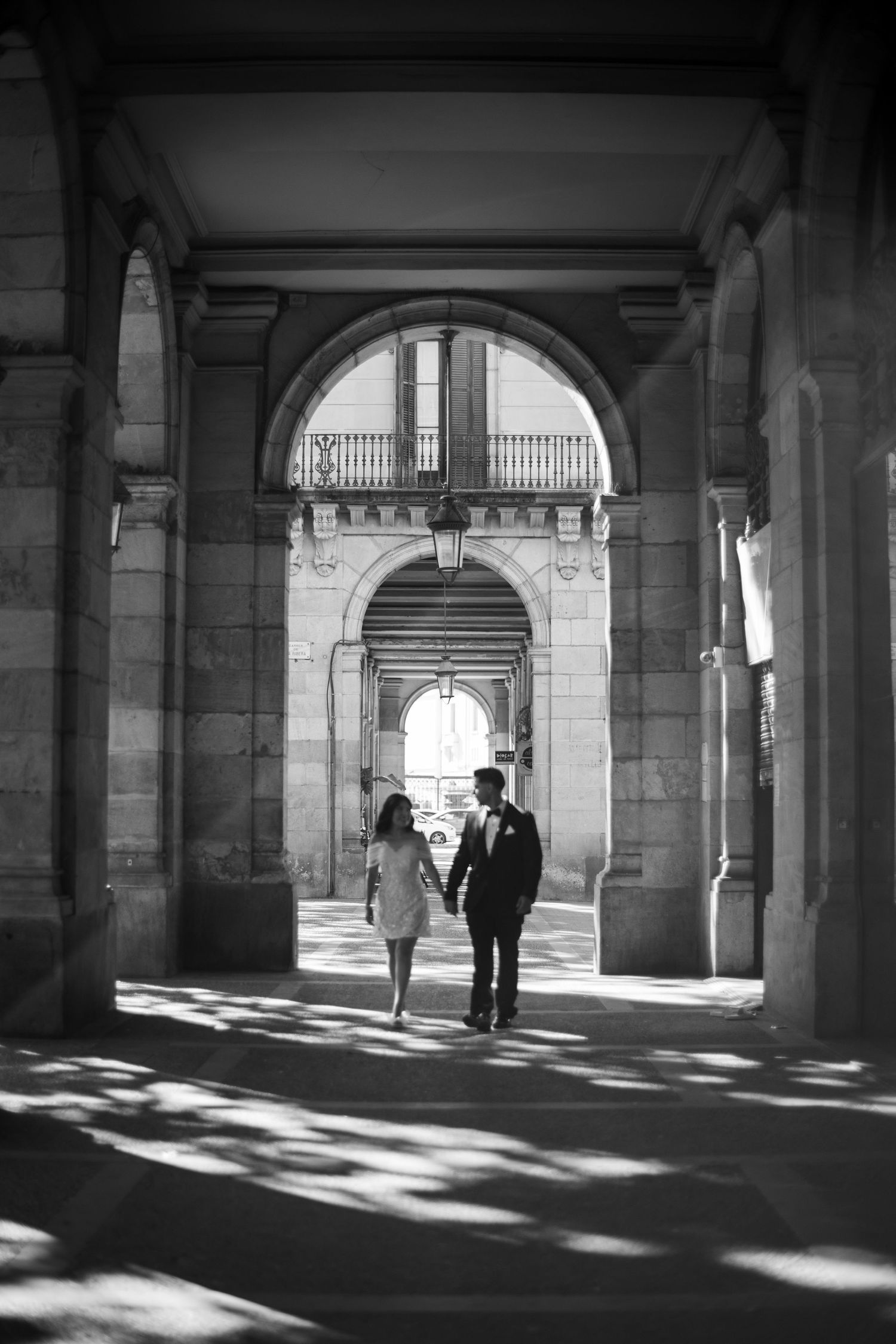 Black and white photo of a couple walking hand in hand through elegant stone archways.