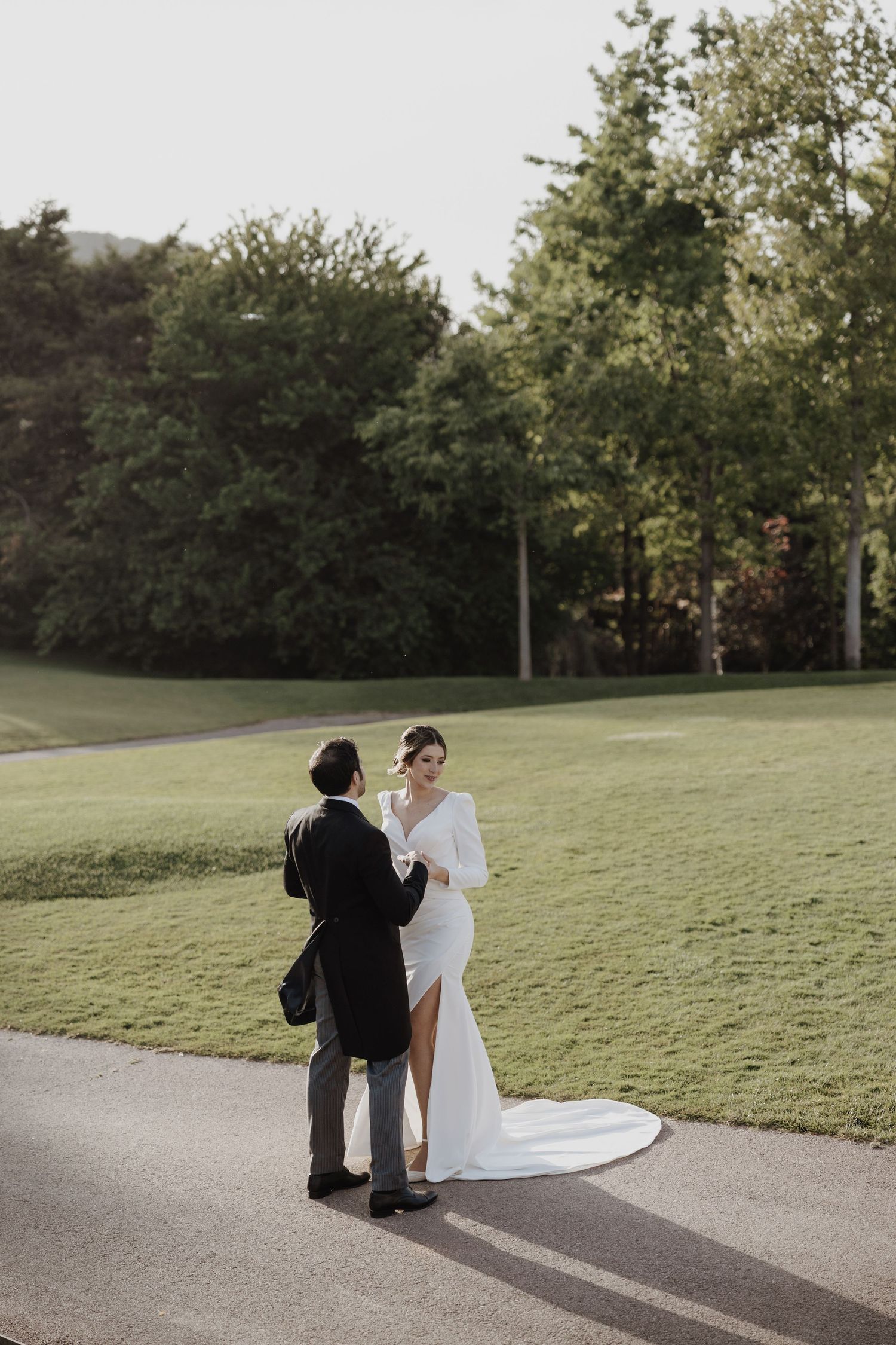 Bride and groom standing on sunlit pathway surrounded by lush green trees and lawn.