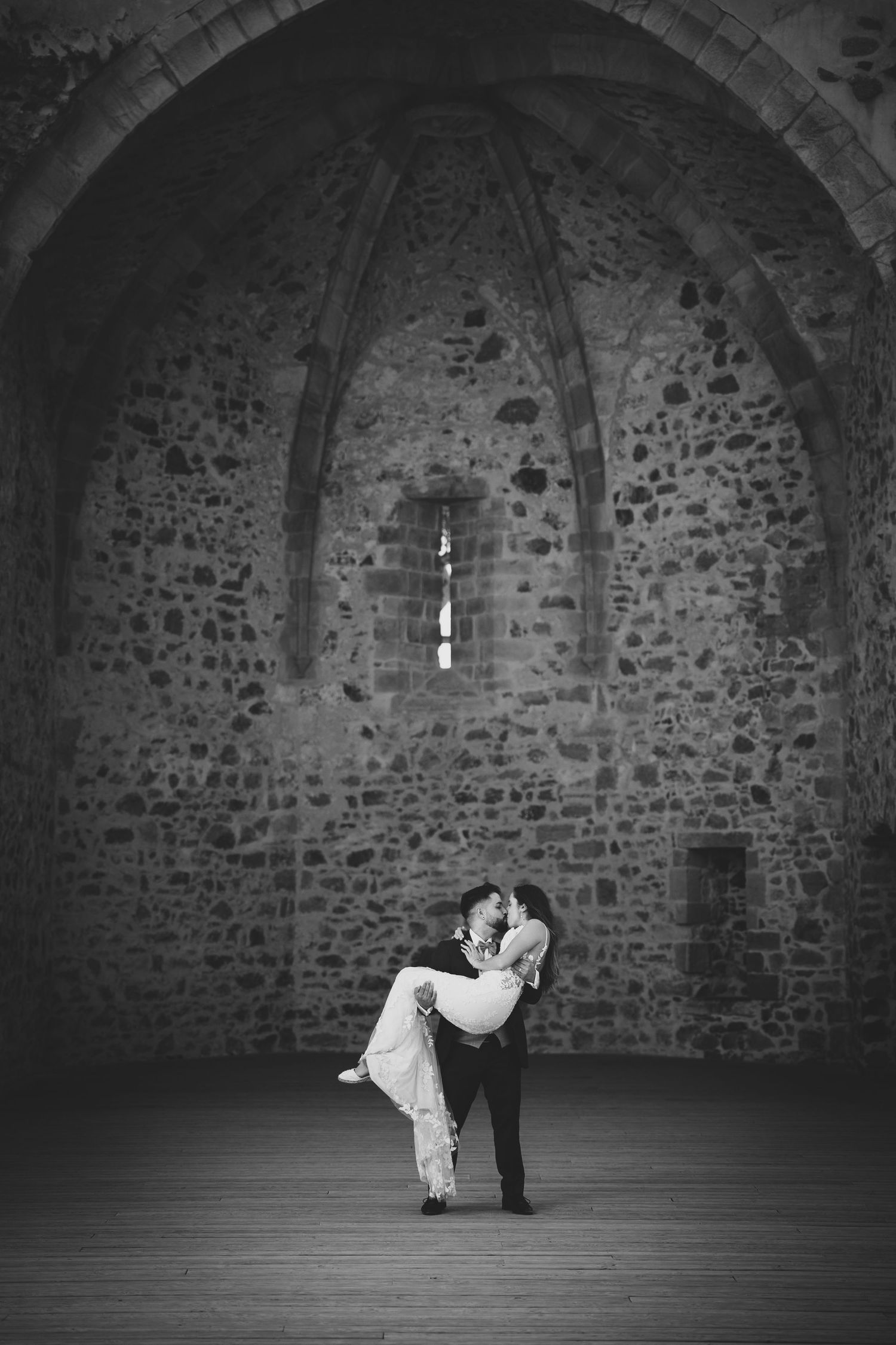 Black and white photo of groom lifting bride in a romantic dip inside a rustic stone arched hall.