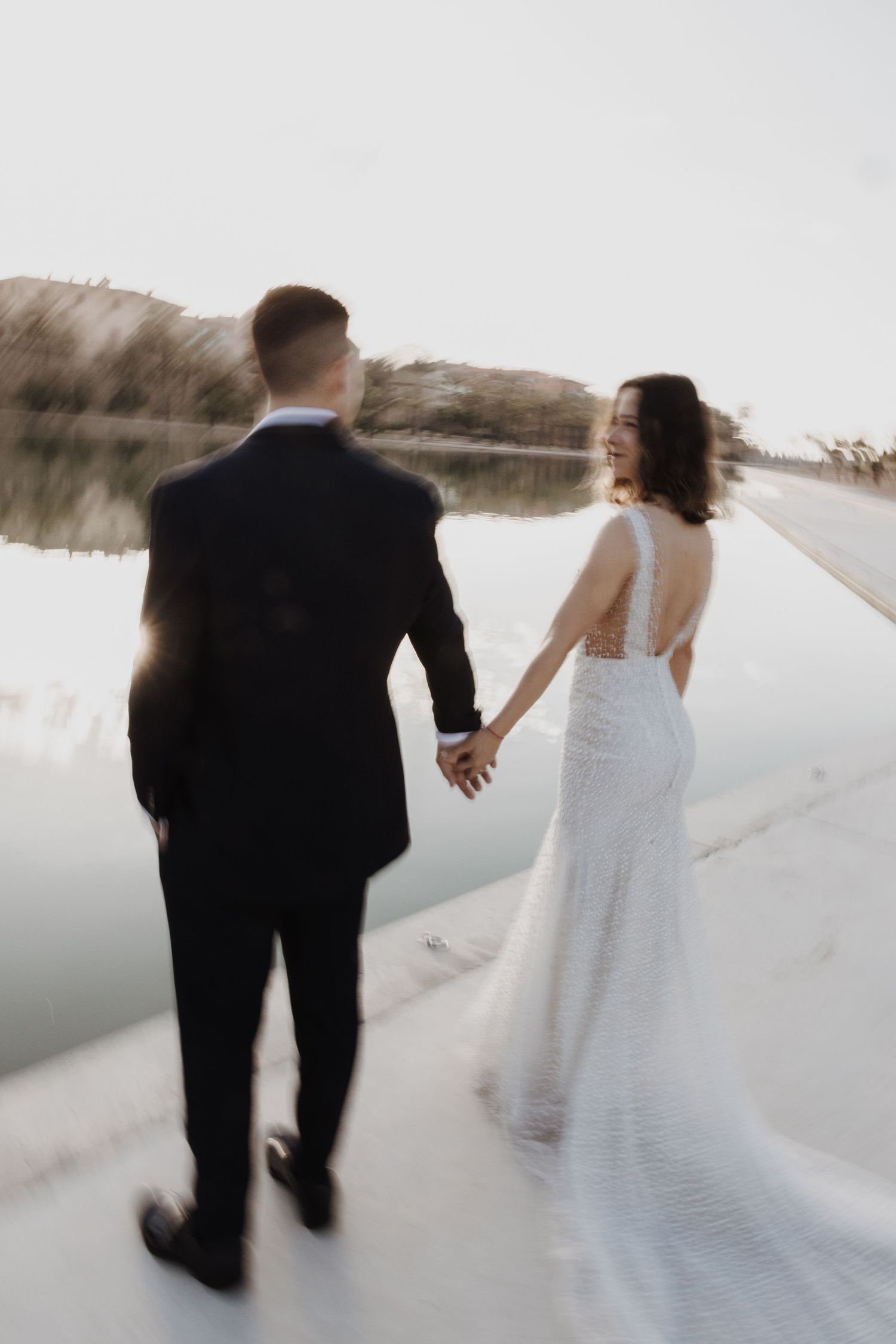 Bride in backless lace gown laughs while holding groom's hand walking near a serene lakeside.