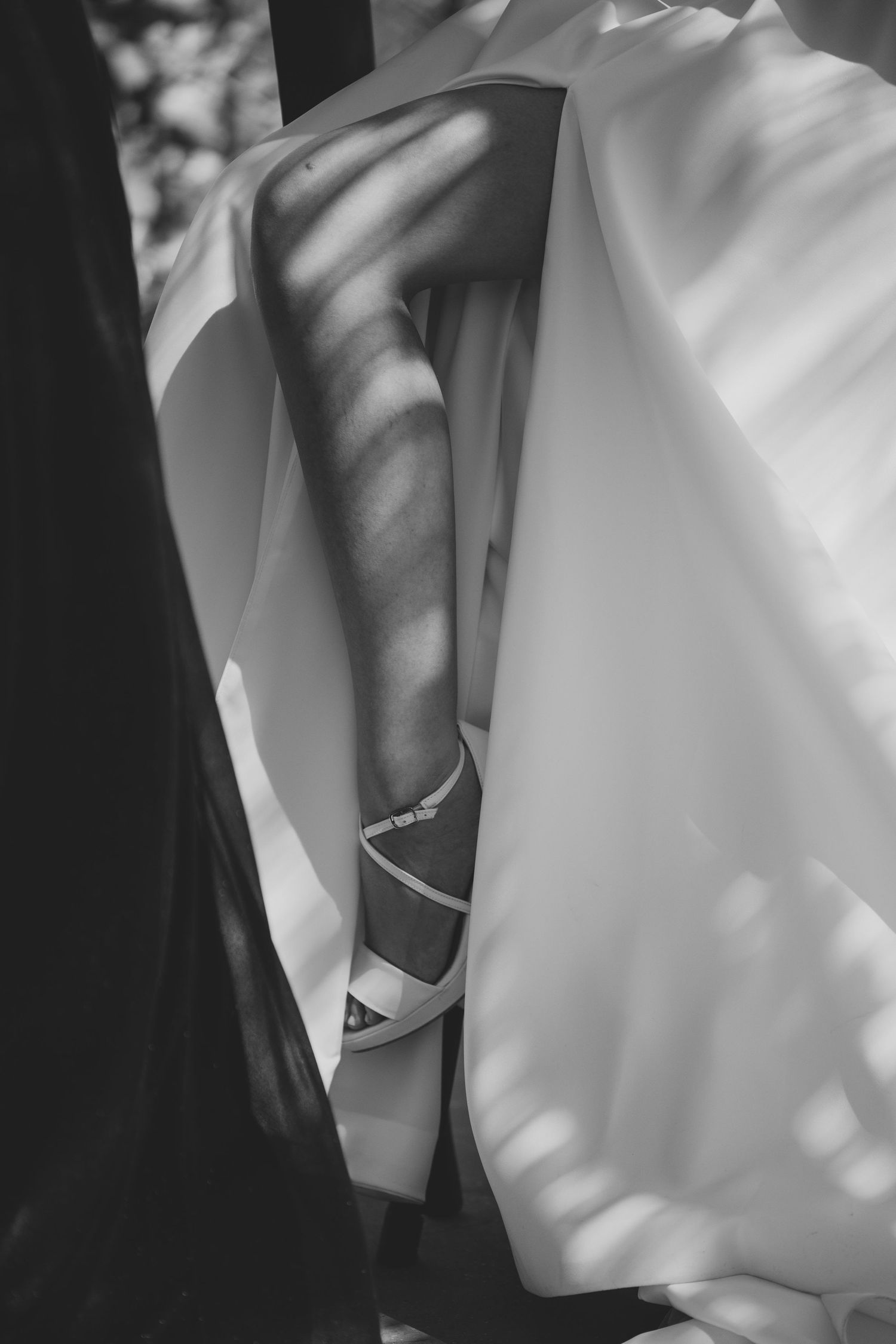 Black and white close-up of a bride's leg in a white strappy heel beneath a flowing wedding dress with dappled sunlight.