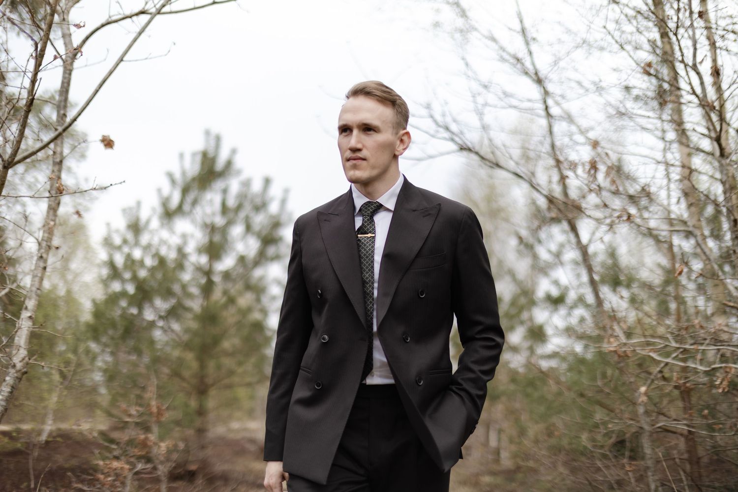 A young man in a dark double-breasted suit and tie stands confidently in a forest setting.