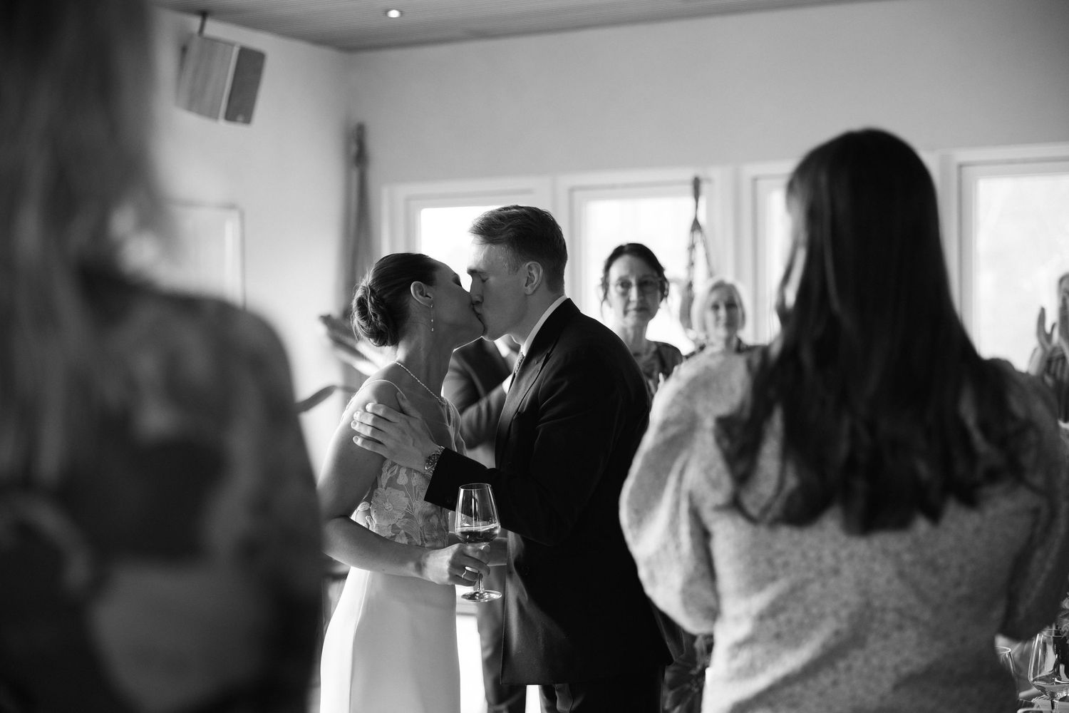 Black and white photo of bride and groom sharing a kiss surrounded by wedding reception guests.