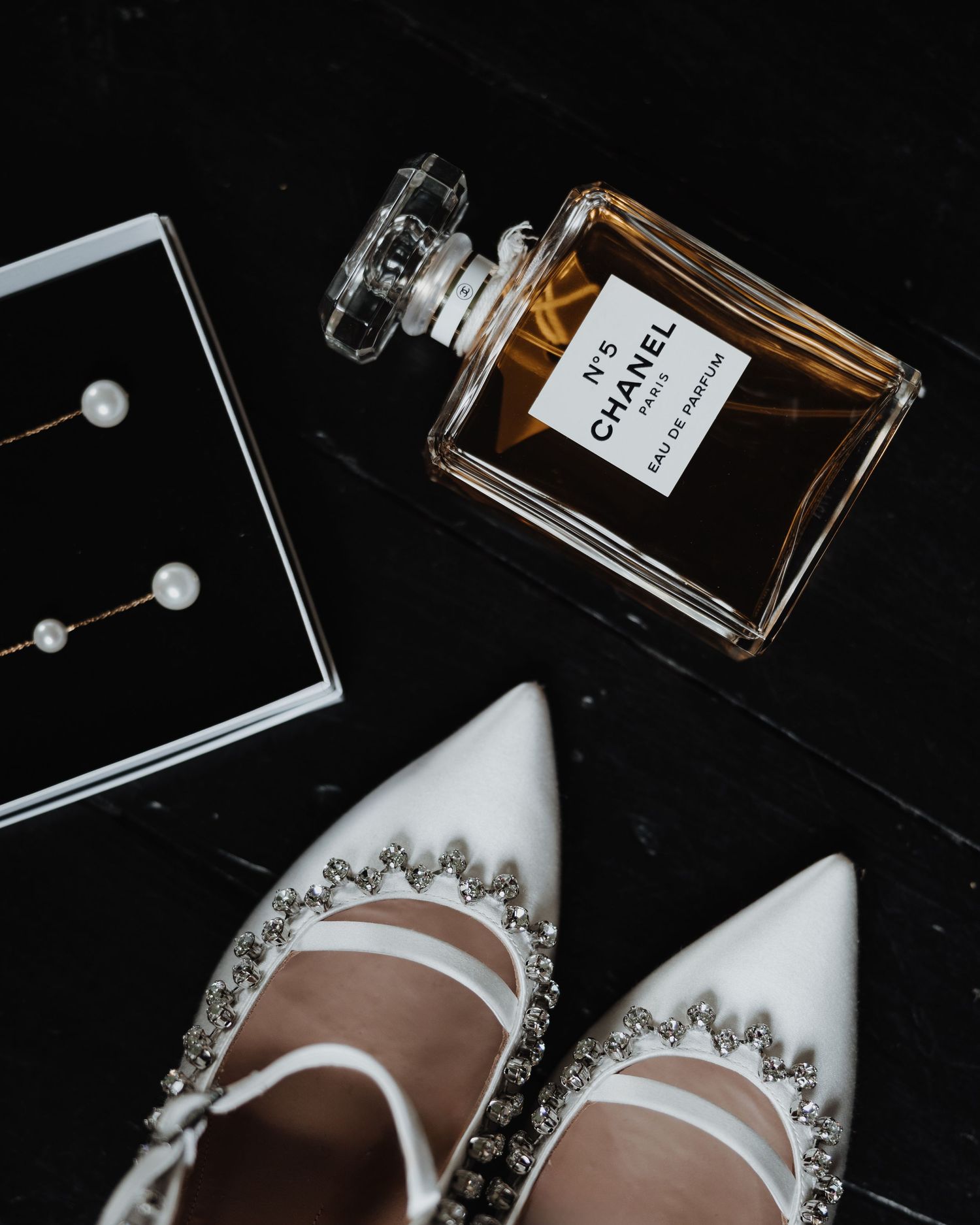 Chanel No.5 perfume bottle with white crystal-embellished bridal heels and pearl drop earrings on dark surface.