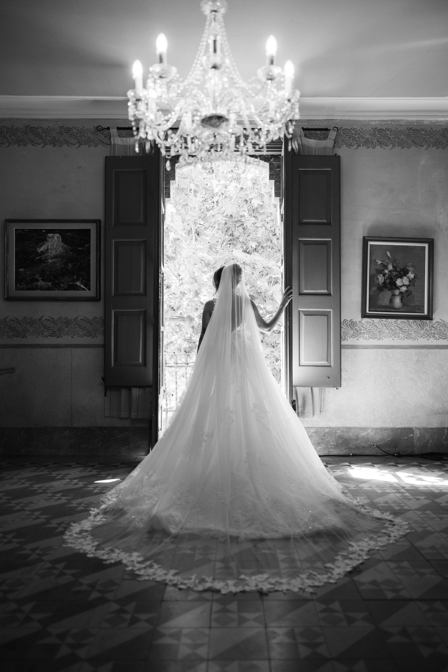A bride in a cathedral veil and ball gown stands before open doors under a crystal chandelier, black and white.