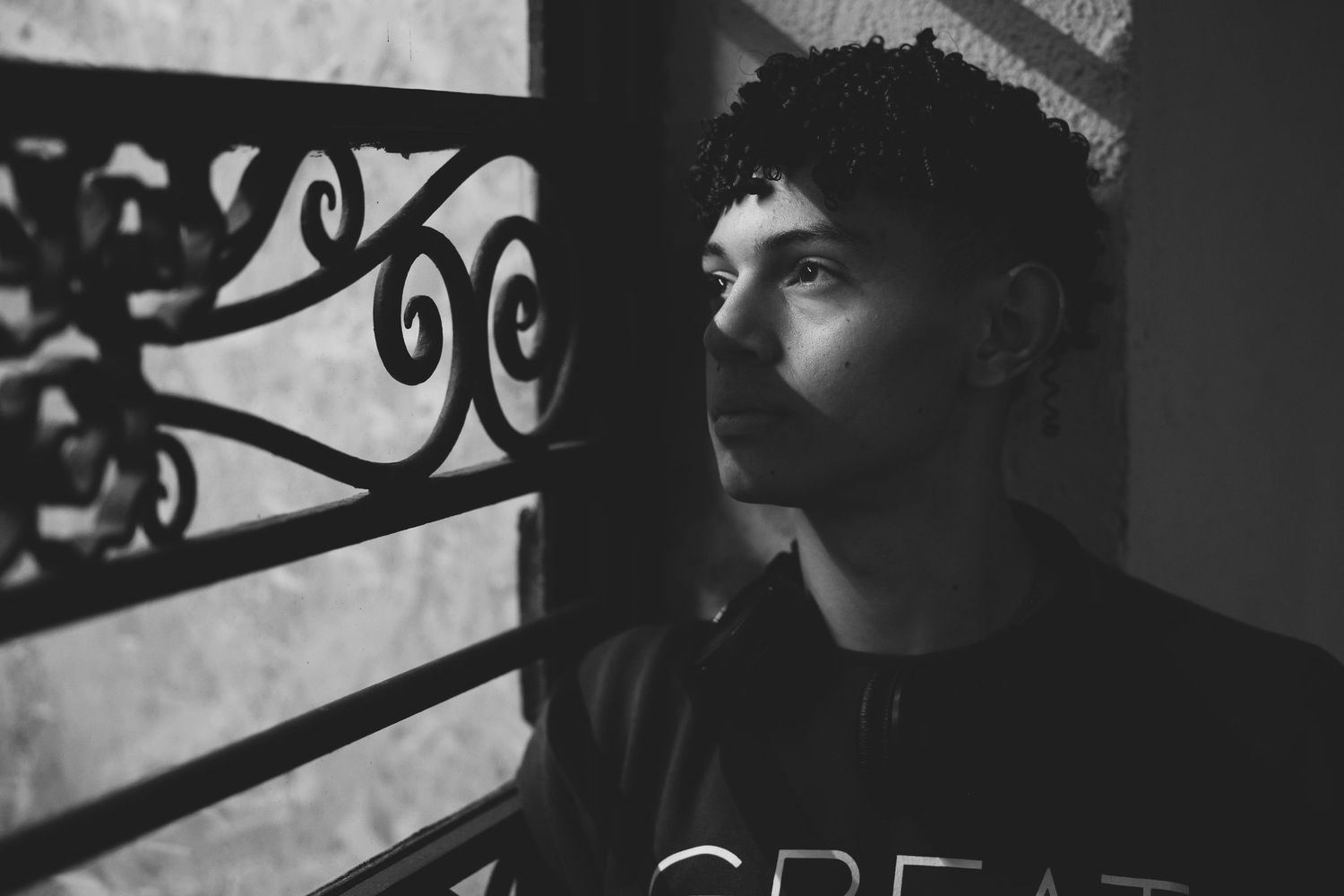 A black and white portrait of a young man with curly hair gazing through an ornate iron gate.