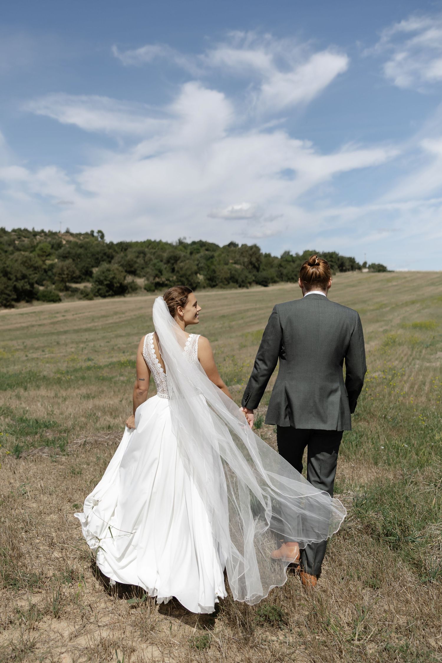 Bride and groom walking hand in hand through an open field with veil flowing in the breeze.