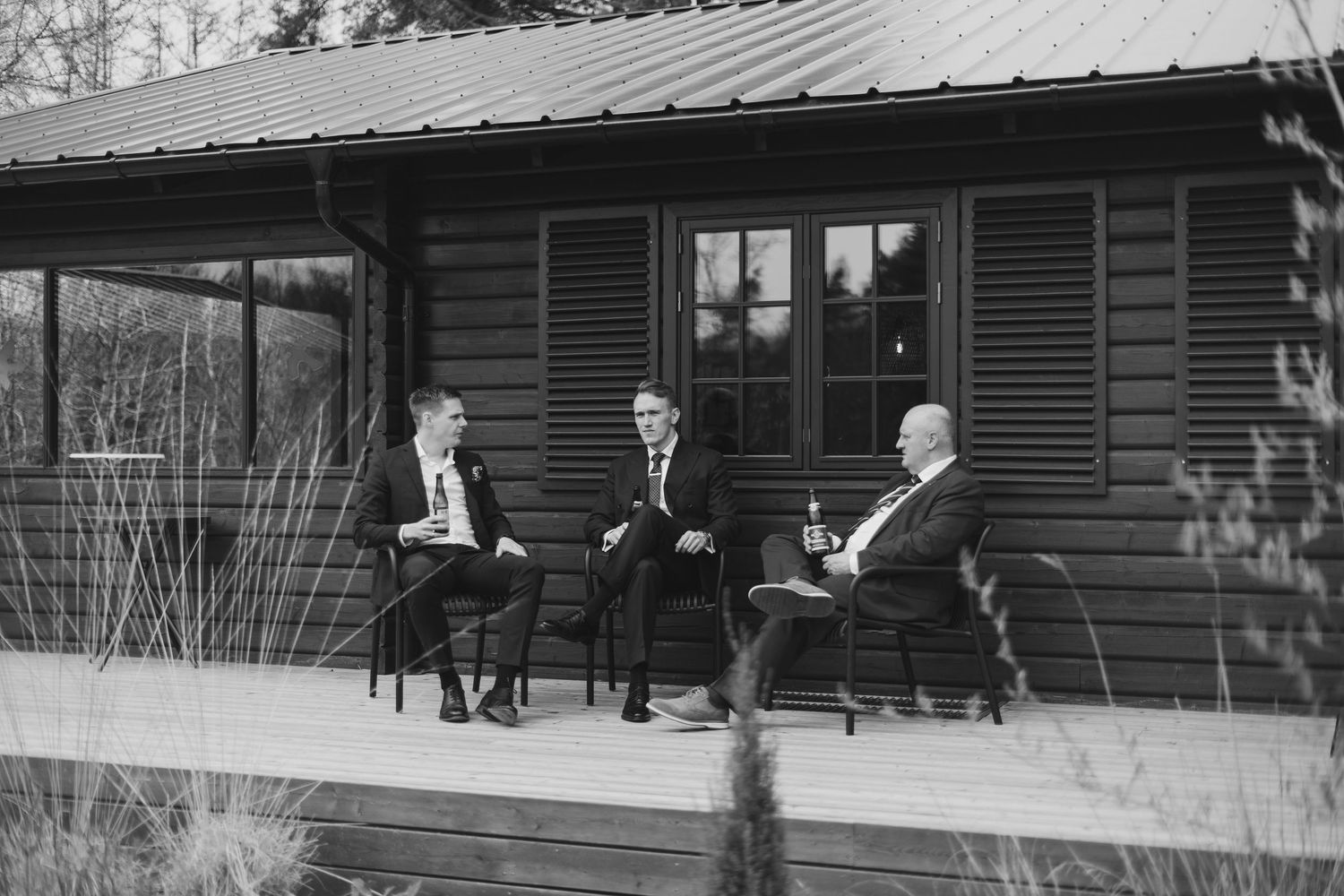Three men in suits sitting on chairs on a dark wooden cabin porch in black and white.
