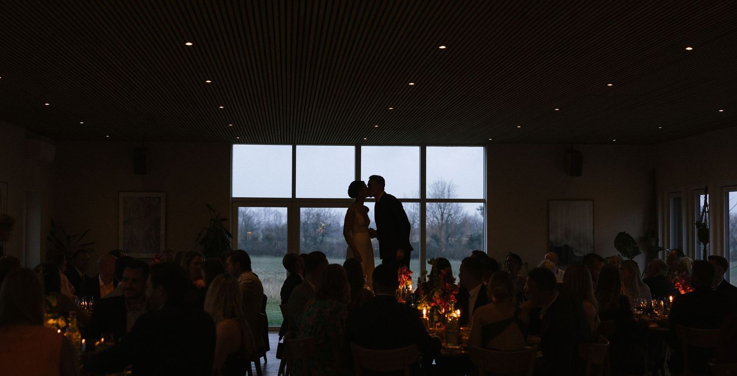 Silhouette of couple kissing at wedding reception with candlelit tables and large windows.