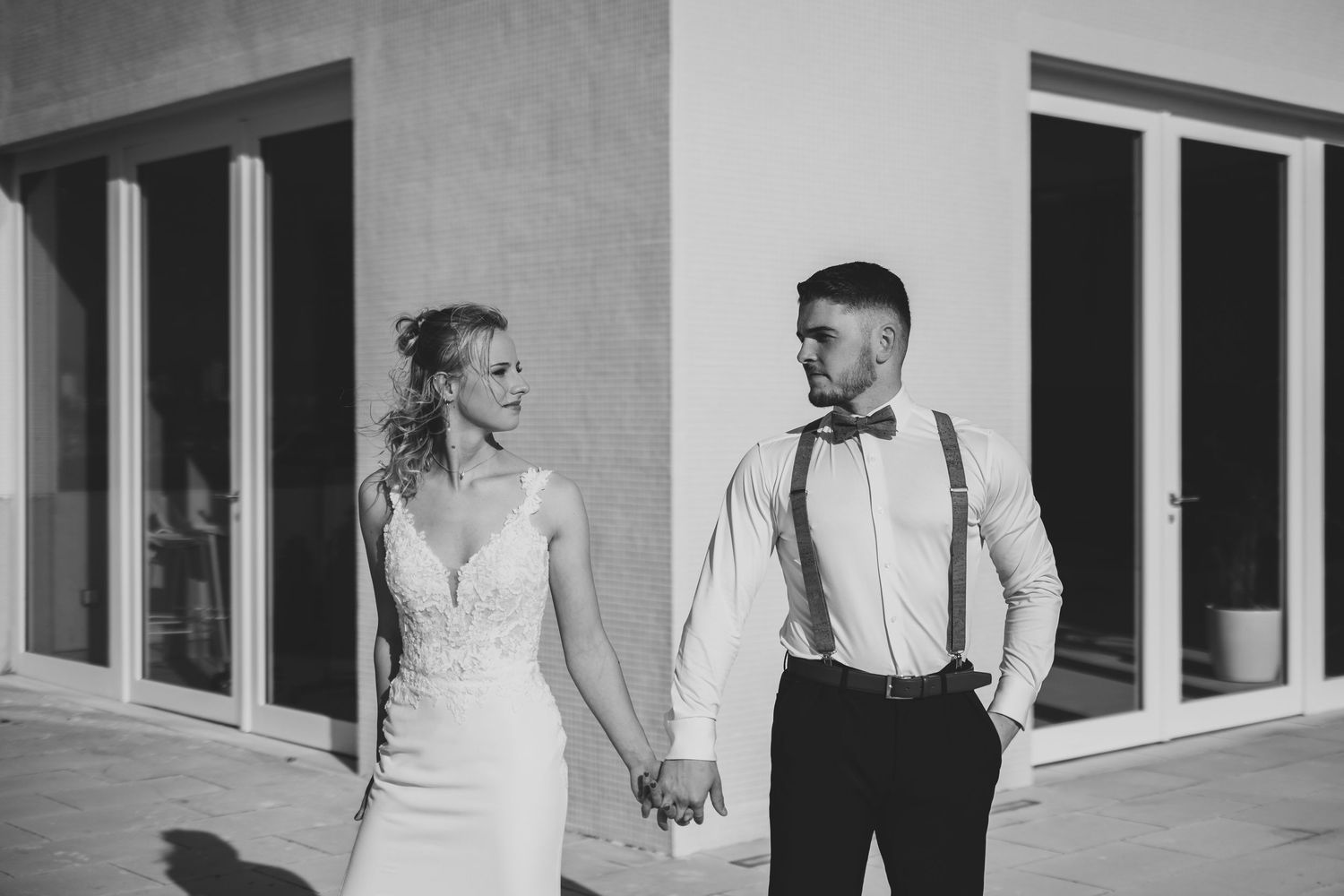 Black and white photo of bride and groom holding hands in front of a modern building exterior.