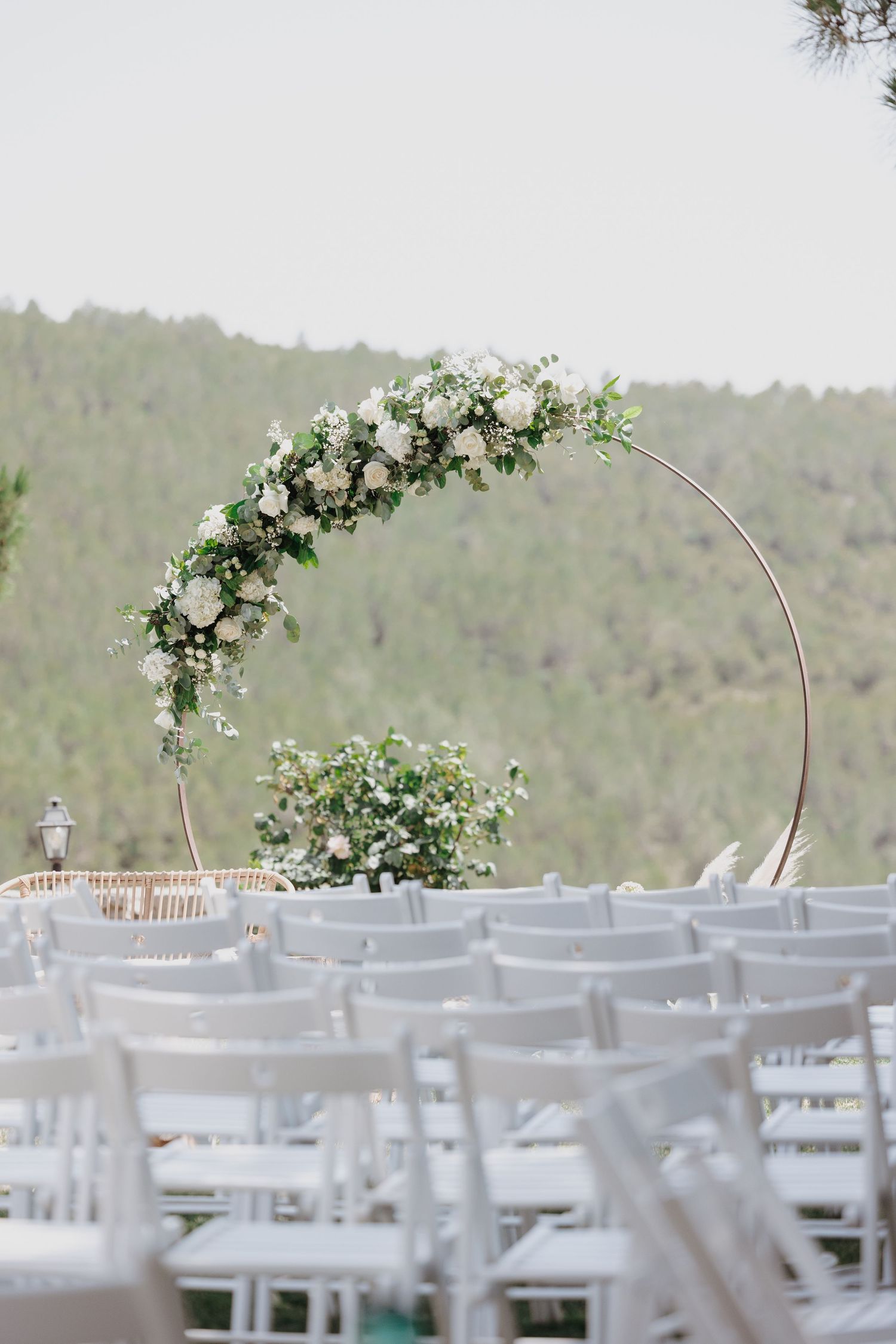 Elegant outdoor wedding ceremony setup with circular floral arch adorned with white flowers and greenery, white chairs arranged in rows.