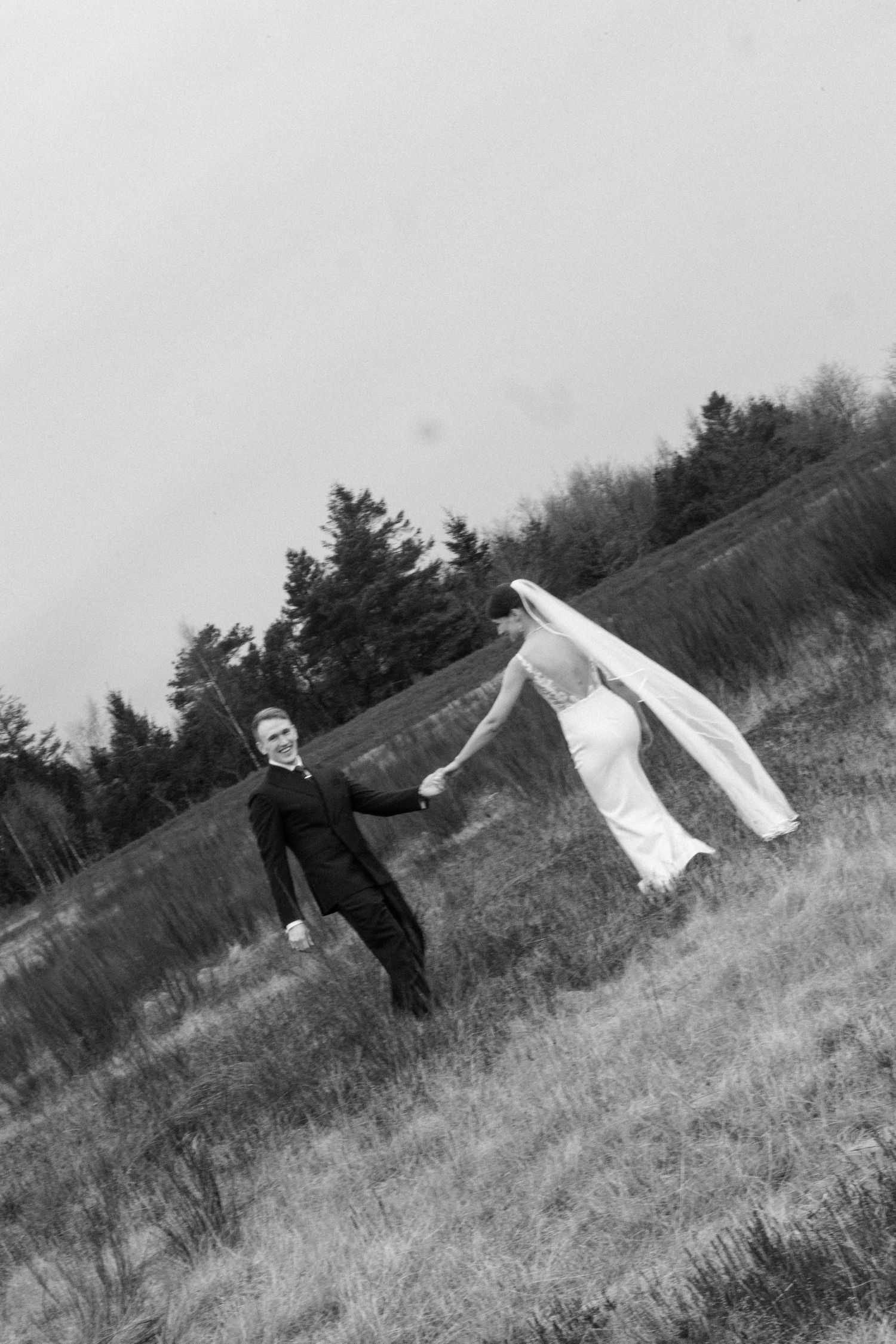 Bride and groom hold hands in a windy field, her veil billowing dramatically in this black and white wedding photo.