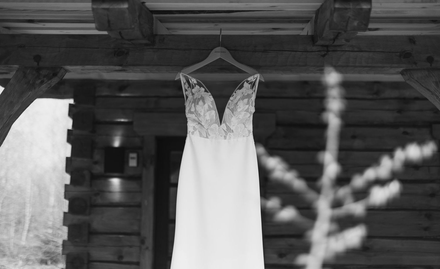 Elegant wedding dress with lace bodice hanging on wooden cabin porch in black and white photo.