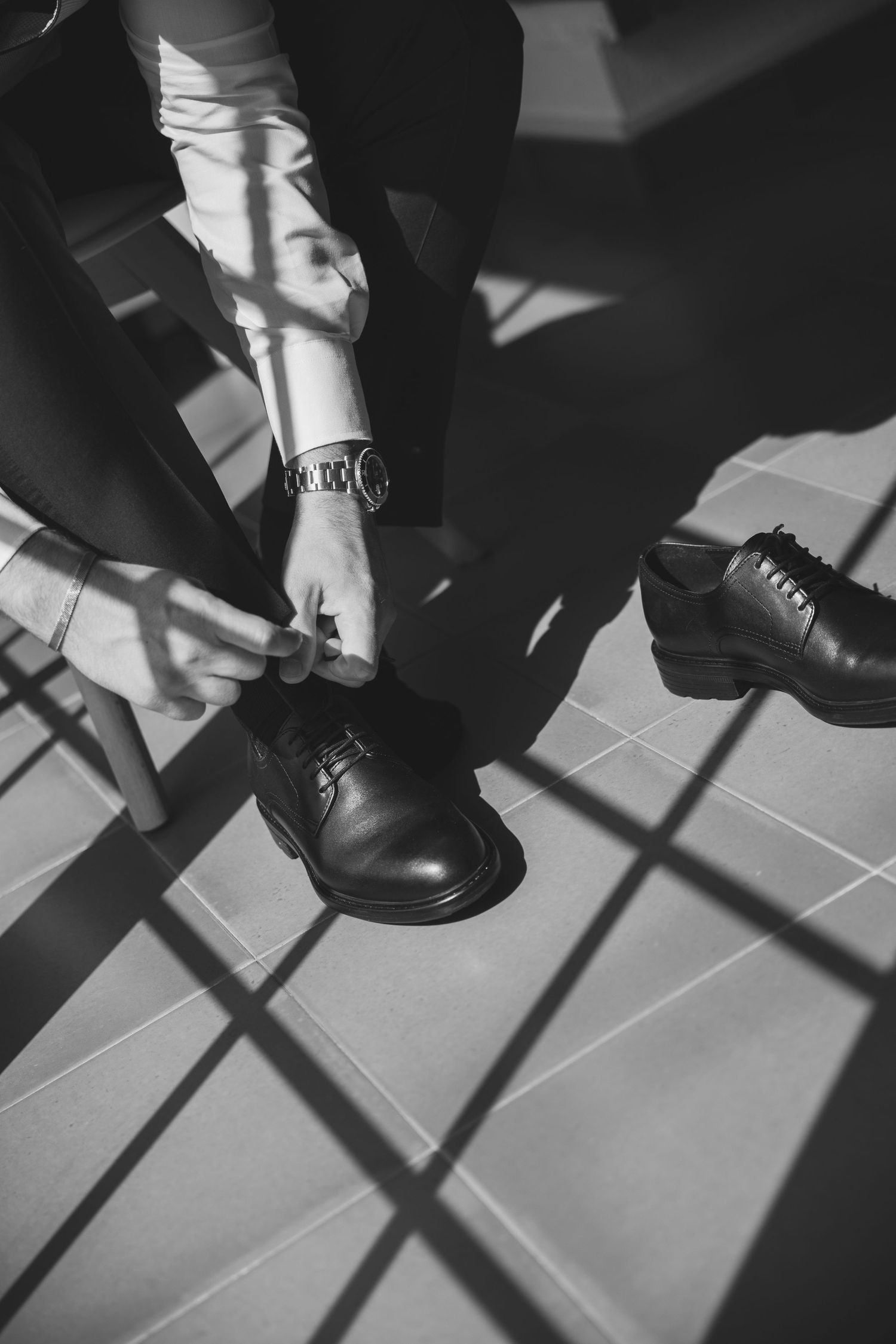Black and white photo of groom lacing up black Oxford dress shoes on tiled floor.
