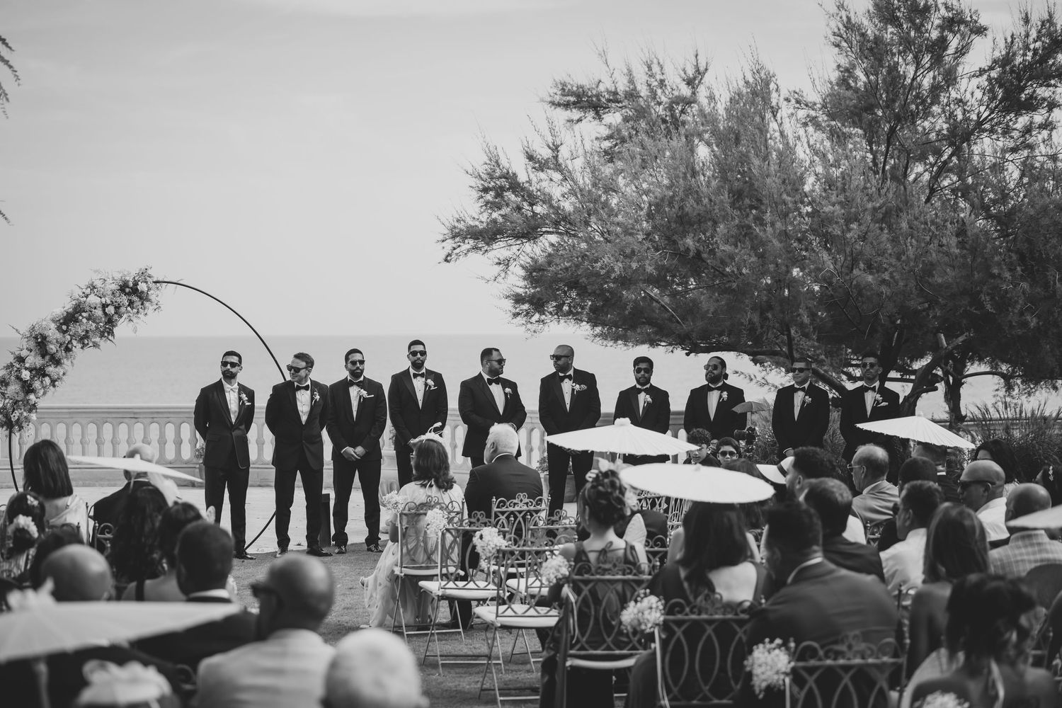 Black and white outdoor wedding ceremony with groomsmen standing at altar overlooking the sea.