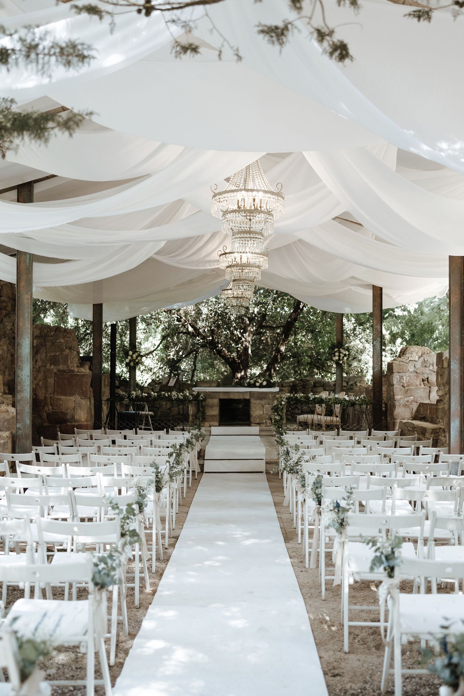 Elegant outdoor wedding ceremony with white draped fabric ceiling, crystal chandelier, white chairs, and a long aisle runner.