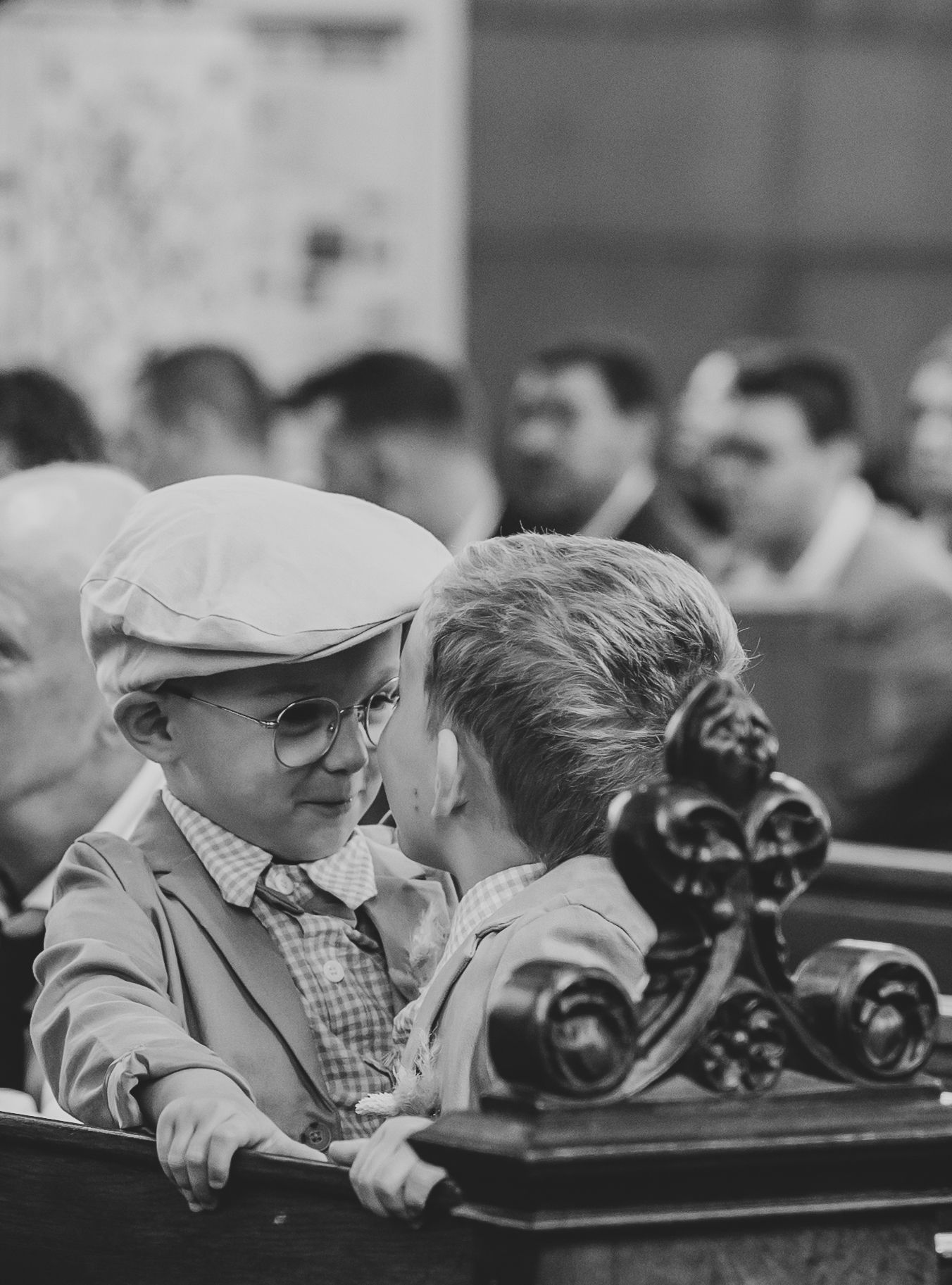 Two young boys in vintage attire sharing a whisper on a church pew during a wedding ceremony.