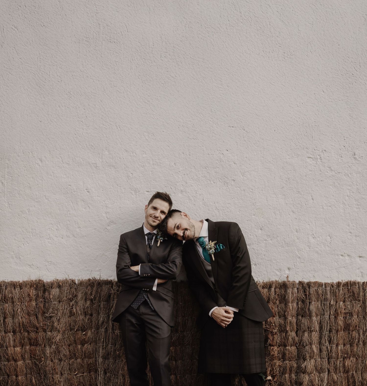 Two grooms in dark suits lean against a white textured wall, smiling warmly on their wedding day.